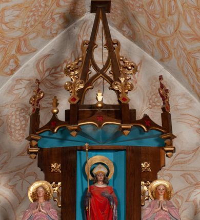 Zdjęcie nr 35: Ołtarz jednokondygnacyjny na wysokim, schodkowym cokole, trójosiowy, z wysuniętą mensą ołtarzową. Płyciny cokołu zdobione dekoracją w formie ostrołukowych maswerków i rozet w kwadratowej ramie. Antepedium flankowane parą kolumn ustawionych na wysokich cokołach i kanelowanych, skręconych trzonach z kompozytowymi kapitelami. W polu dwie prostokątne płyciny i kwadratowa pośrodku ze stylizowaną wicią roślinną i małą uskrzydloną główką aniołka. Tabernakulum proste, w kształcie prostopadłościennej skrzynki, o złotych drzwiczkach z wytłaczaną formą równoramiennego krzyża i wypukłą srebrną rybą oraz koszem z chlebem. Po obu stronach, na niewysokich postumentach dwa klęczące anioły z wysokimi skrzydłami, w biało różowych szatach z fioletowym podbiciem (skrzydła stylizowane na husarskie). Anioł po lewej z kadzielnicą, a anioł po prawej z kielichem.
Nastawa ołtarzowa o trzech oddzielnych niszach, nakrytych gotycyzującymi baldachimami z pinaklami, ozdobionymi żabkami. Oś środkowa w formie niszy zamkniętej ostrołukowo z wimpergą wspartą na smukłych, złoconych kolumienkach o kapitelach kompozytowych. Wimperga ze złoconymi, profilowanymi płycinami, zwieńczona rzeźbionymi liśćmi akantu i wolutami. Na przedłużeniu kolumn postumenty z figurami aniołów w biało niebieskich szatach, ze złotymi, wydłużonymi skrzydłami. Na osi na postumencie, pod gotycyzującym baldachimem figura świętego biskupa w czerwonej szacie z krzyżem w dłoni pomiędzy dwoma aniołami w białych i różowych szatach. W niszy, na czerwonym tle obraz Matki Boskiej Nieustającej Pomocy w prostej ramie z dekoracją w formie wici roślinnej. Poniżej dwa płaskorzeźbione anioły trzymające ażurowy monogram maryjny. Anioły o pozłacanych skrzydłach, na chmurach, w różowej i niebieskiej szacie. Osie boczne w formie aediculi z gotycyzującym baldachimem. W niszach figury, po lewej św. Piotra, a po prawej św. Pawła. Aedicule ujęte są smukłymi, skręconymi kolumnami z kapitelami wolutowymi oraz roślinnym uszakiem z perełkowaniem. Nad nimi imposty, na których rzeźby osłaniane baldachimami, po lewej świętego w habicie jezuity, a po prawej św. Jana Kantego. Drewno bejcowane na ciemny brąz, z nakładanymi elementami dekoracyjnymi złoconymi. 