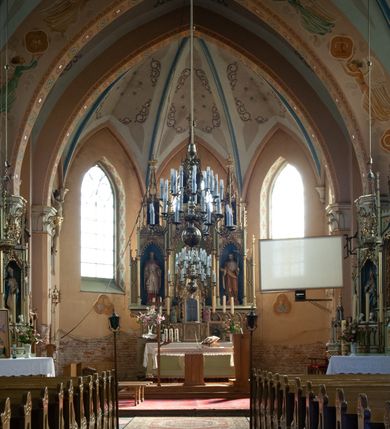 Zdjęcie nr 31: Kościół ceglany, na planie prostokąta z węższym i niższym prezbiterium z zakrystią od północy i wieżą od zachodu. Kościół orientowany, jednonawowy, czteroprzęsłowy z węższym, jednoprzęsłowym prezbiterium zamkniętym trójbocznie, z kruchtą w przyziemiu wieży. Nawa artykułowana półfilarami o ściętych narożach, przechodzącymi w zamknięte łukiem ostrym gurty krzyżowo-żebrowego sklepienia. W nawie i prezbiterium okna prostokątne zamknięte łukiem ostrym – pięć w prezbiterium, po trzy z każdej strony nawy. Węższe okna w przęśle chóru, kolejne w fasadzie. Główne wejście od wschodu, drugie od zachodu. Chór muzyczny w czwartym przęśle nawy, wsparty na wielobocznych filarach, podtrzymujących ostrołukowe arkady, z parapetem ozdobionym blendami. Ściany w dolnej partii pokryte drewnianą boazerią, wyżej malowane na kremowo, w partiach sklepienia polichromia podkreślająca elementy konstrukcji fragmentami stylizowanej wici i dekoracji floralnej; na belce tęczowej anioły niosące plakiety z symbolami świętych sakramentów. Fasada trójosiowa, z ryzalitem szerokości wieży oszkarpowanym na narożach. Podziały poziome przeprowadzone przy pomocy gzymsów. Na osi ostrołukowe wejście, powyżej wypełniony maswerkiem okulus. Następna kondygnacja jest częścią czworobocznej wieży, wypełniono ją dużym oknem w kształcę stojącego prostokąta zamkniętego łukiem ostrym.  Boczne osie z małymi oknami powtarzającymi kształt pozostałych, zamknięte trójkątnym przyczółkiem przerwanym na wieżę. Najwyższa kondygnacja wieży przepruta z każdej strony parą okien w kształcie stojących prostokątów umieszczonych w ostrołukowych niszach. Wieńczący ją gzyms dekorowany zygzakiem, powyżej trójkątne szczyty z ostrołukową blendą z okulusem w centrum. Elewacje boczne i prezbiterium oszkarpowane, przeprute oknami.  Hełm wieży w formie latarni zakończonej stożkiem z kupą i krzyżem, nawa przekryta dachem dwuspadowym, prezbiterium wielobocznym, zakrystia i wejście od zachodu pulpitowymi. 


Do najciekawszych elementów wyposażenia należą zespół neogotyckich ołtarzy wraz z amboną oraz kolekcja dziewiętnastowiecznych feretronów. W otoczeniu kościoła znajduje się kamienna figura św. Jana Nepomucena z 1788 roku. 

