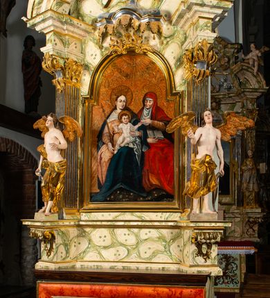 Zdjęcie nr 2: Ołtarz architektoniczny, jednoosiowy, jednokondygnacyjny, ze zwieńczeniem. Ołtarz prostopadłościenny z drewnianą, współczesną mensą i współczesnym antependium z przedstawieniem świętej w otoczeniu wieńca z kwiatami. Nastawa ujęta ukośnie dostawionymi filarami kanelowanymi, ustawionymi na postumentach z konsolami o prostych kapitelach utworzonych z roccailli i konsol podtrzymujących przerwane belkowanie zwieńczone ćwierćkolistymi naczółkami. Zwieńczenie o wklęsłych bokach zakończone profilowanym gzymsem w kształcie łuku nadwieszonego półkolistego. Na tle filaru, na konsolach ustawione figury uskrzydlonych aniołów. W polu środkowym obraz w kształcie stojącego prostokąta zamkniętego łukiem nadwieszonym półkolistym ozdobiony rocaillem. W ramie obraz z przedstawieniem św. Anny Samotrzeciej z Madonną z Dzieciątkiem. Nad ramą baldachim z lambrekinem. W zwieńczeniu serce Gorejące na tle glorii promienistej z chmurkami, zwieńczonej uskrzydloną główką anielską; nad gzymsem zwieńczenia  grzebień utworzony z ażurowego rocaillu. 