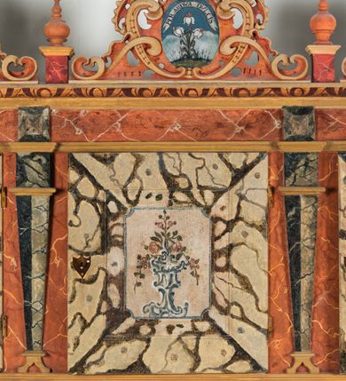 Zdjęcie nr 59: Umeblowanie zakrystii usytuowane jest wzdłuż ścian północnej, południowej i wschodniej pomieszczenia.  Na ścianie północnej, w części dolnej znajdują się trzy połączone ze sobą komody, każda o czterech szufladach, nakryte jednym blatem. W części górnej mieszczą się pięcioosiowe szafki z szufladami w cokole. Osie artykułowe są pilastrami hermowymi. Na osi środkowej szafka jest nieco wyższa i szersza z drzwiczkami dekorowanymi prostokątną, uszatą, oprofilowaną płyciną z malowanym przedstawieniem Chrystusa Eucharystycznego. Drzwiczki pozostałych szafek mają kształt stojących prostokątów i dekorowane są płycinami o wklęsłych narożach, zdobionymi gałązkami czerwonych róż, ułożonych w rokokowych wazonach. Ścianki zamknięte są niepełnym belkowaniem z gzymsem. W partii zwieńczenia osie artykułowane są cokolikami, na których ustawione są toczone, kuliste wazoniki. Pomiędzy nimi umieszczone są kartusze utworzone z ornamentu małżowinowo-chrząstkowego z malowanymi emblematami na owalnych medalionach, które ułożone są kolejno od lewej strony: 
1. Pionowo ustawiony miecz, ostrzem skierowany ku górze, połączony ze skrzyżowanymi berłem i dwoma kluczami, od których odchodzą fantazyjnie ułożone sznury w kolorze czerwonym. U góry napis na szarfie: „OBEDITE”.
2. W górnej części wyłaniająca się z chmur ręka trzymająca w dłoni węgielnicę i przymocowane do niej na łańcuszku tablice Dekalogu. Napis na szarfie: „OMNIA SVSTINET”.
3. Bóg Ojciec ukazany w półpostaci, lewą ręką opiera się na kuli ziemskiej, prawą czyni gest błogosławieństwa. 
4. Bujnie ulistnione, ciemnozielone drzewko spięte w dolnej części złotą formą w kształcie dwóch wywiniętych wolutek. Napis na szarfie: „GAVDET PATIENTIA DVRIS”. 
5. W górnej części obrazu z chmur wyłania się ręka, która zrzuca na ziemię iskrzący pocisk i dwa skrzyżowane pioruny, połączone parą białych skrzydeł. Napis „FORTIS VLTOR”.
 
Na ścianie środkowej, wschodniej znajduje się sarkofagowa mensa, powyżej parawanowa ścianka na profilowanym cokole, artykułowana pilastrami, pomiędzy którymi umieszczone są płyciny z kształcie leżących prostokątów z obrazami; ścianka zamknięta jest profilowanym gzymsem. W zwieńczeniu, na jej skrajach znajdują się owalne medaliony ujęte w kartusze z ornamentu małżowinowo-chrząstkowego. W medalionie po lewej stronie namalowane są litery alfa i omega na błękitnym tle oraz wstęga z napisem: „RENOVATUM A(NNO) D(OMINI) 1975”. Po prawej stronie analogiczny kartusz z literami w medalionie: „Ω / O / N” wpisane w formę krzyża, otoczonego niepełną aureolą; powyżej wstęga z napisem: „VIDENTEM AUDERE”. W płycinach obrazy przedstawiają świętych w ujęciu do pasa, zwróconych ku środkowi; od lewej:
1. Święty Demetriusz z włócznią opartą o prawy bark, ubrany w habit zakonu kanoników laterańskich od pokuty.
2. Święty Klet z aniołkiem, który trzyma w dłoni palmę męczeństwa i krzyż papieski. W prawym górnym narożu obrazu napis: „FAC HVIC SIGNO HONOREM”.
3. Święty Augustyn z gorejącym sercem na piersi i aniołkiem z łyżką i miseczką w dłoniach.
4. Święty ubrany w habit Marków, który trzyma w dłoni serce z krzyżem, podtrzymywane przez Chrystusa na obłokach – prawdopodobnie św. Cyriak, założyciel zakonu.
 
Na ścianie południowej znajduje się analogiczne umeblowanie, jak na ścianie północnej. W dolnej części dwie komody, a w miejscu trzeciej od wschodu szafka zdobiona dwiema uszatymi płycinami. Górna część mebla w przeciwieństwie do ściany północnej bez podwyższenia części środkowej. 
Na ścianie południowej:
1. Jasnobrązowa harfa na błękitnym tle i napis: „IN DOMINO”.
2. Ciemnobrązowy ptak i trzy podrywające się do lotu młode, w górnej części złote słońce i napis: „NON QUAE SVPER TERRAM.”
3. Po lewej stronie kompozycji wyłaniająca się z chmur ręka trzymająca skrzyżowane: miecz, czerwony krzyż i palmę. Napis: „NON ERVBESCO”.
4. W centrum przedstawienia krzak białej lilii o trzech rozgałęzieniach, w tle chmury w odcieniach koloru szaroniebieskiego. Napis na szarfie: „PER ARDVA DVLCIS”.
5. W lewej części kompozycji uskrzydlony jeleń, wspięty na tylnych nogach, po prawej stronie czarne i brązowe skały, w partii górnej otwarta korona, umieszczona pośrodku obłoku obwiedzionego biało-czerwonym pasem. Napis: „ET COMPREHENDATIS”.
 
W miejscu połączenia komód ścian północnej i południowej z dolną częścią konstrukcji na ścianie wschodniej znajdują się ukośnie ustawione, jednoskrzydłowe szafki o drzwiczkach dekorowanych prostokątną płyciną o wklęsłych narożach. Ścianki boczne części północnej i południowej zdobione są ukośną kratką. Powierzchnia szafek i komód pokryta jest polichromią imitującą marmur w kolorze beżowym, szarokremowym, różowym i rdzawym. Blaty malowane są farbą olejną w kolorze beżowym. Obramienia medalionów malowane w kolorze rdzawozłotym. Tło w większości medalionów w odcieniach błękitu, w górnych częściach majuskułowe napisy objaśniające przedstawienie, umieszczone w białych i półkoliście ułożonych szarfach. 