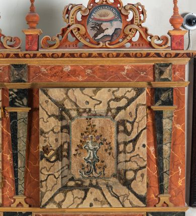 Zdjęcie nr 69: Umeblowanie zakrystii usytuowane jest wzdłuż ścian północnej, południowej i wschodniej pomieszczenia.  Na ścianie północnej, w części dolnej znajdują się trzy połączone ze sobą komody, każda o czterech szufladach, nakryte jednym blatem. W części górnej mieszczą się pięcioosiowe szafki z szufladami w cokole. Osie artykułowe są pilastrami hermowymi. Na osi środkowej szafka jest nieco wyższa i szersza z drzwiczkami dekorowanymi prostokątną, uszatą, oprofilowaną płyciną z malowanym przedstawieniem Chrystusa Eucharystycznego. Drzwiczki pozostałych szafek mają kształt stojących prostokątów i dekorowane są płycinami o wklęsłych narożach, zdobionymi gałązkami czerwonych róż, ułożonych w rokokowych wazonach. Ścianki zamknięte są niepełnym belkowaniem z gzymsem. W partii zwieńczenia osie artykułowane są cokolikami, na których ustawione są toczone, kuliste wazoniki. Pomiędzy nimi umieszczone są kartusze utworzone z ornamentu małżowinowo-chrząstkowego z malowanymi emblematami na owalnych medalionach, które ułożone są kolejno od lewej strony: 
1. Pionowo ustawiony miecz, ostrzem skierowany ku górze, połączony ze skrzyżowanymi berłem i dwoma kluczami, od których odchodzą fantazyjnie ułożone sznury w kolorze czerwonym. U góry napis na szarfie: „OBEDITE”.
2. W górnej części wyłaniająca się z chmur ręka trzymająca w dłoni węgielnicę i przymocowane do niej na łańcuszku tablice Dekalogu. Napis na szarfie: „OMNIA SVSTINET”.
3. Bóg Ojciec ukazany w półpostaci, lewą ręką opiera się na kuli ziemskiej, prawą czyni gest błogosławieństwa. 
4. Bujnie ulistnione, ciemnozielone drzewko spięte w dolnej części złotą formą w kształcie dwóch wywiniętych wolutek. Napis na szarfie: „GAVDET PATIENTIA DVRIS”. 
5. W górnej części obrazu z chmur wyłania się ręka, która zrzuca na ziemię iskrzący pocisk i dwa skrzyżowane pioruny, połączone parą białych skrzydeł. Napis „FORTIS VLTOR”.
 
Na ścianie środkowej, wschodniej znajduje się sarkofagowa mensa, powyżej parawanowa ścianka na profilowanym cokole, artykułowana pilastrami, pomiędzy którymi umieszczone są płyciny z kształcie leżących prostokątów z obrazami; ścianka zamknięta jest profilowanym gzymsem. W zwieńczeniu, na jej skrajach znajdują się owalne medaliony ujęte w kartusze z ornamentu małżowinowo-chrząstkowego. W medalionie po lewej stronie namalowane są litery alfa i omega na błękitnym tle oraz wstęga z napisem: „RENOVATUM A(NNO) D(OMINI) 1975”. Po prawej stronie analogiczny kartusz z literami w medalionie: „Ω / O / N” wpisane w formę krzyża, otoczonego niepełną aureolą; powyżej wstęga z napisem: „VIDENTEM AUDERE”. W płycinach obrazy przedstawiają świętych w ujęciu do pasa, zwróconych ku środkowi; od lewej:
1. Święty Demetriusz z włócznią opartą o prawy bark, ubrany w habit zakonu kanoników laterańskich od pokuty.
2. Święty Klet z aniołkiem, który trzyma w dłoni palmę męczeństwa i krzyż papieski. W prawym górnym narożu obrazu napis: „FAC HVIC SIGNO HONOREM”.
3. Święty Augustyn z gorejącym sercem na piersi i aniołkiem z łyżką i miseczką w dłoniach.
4. Święty ubrany w habit Marków, który trzyma w dłoni serce z krzyżem, podtrzymywane przez Chrystusa na obłokach – prawdopodobnie św. Cyriak, założyciel zakonu.
 
Na ścianie południowej znajduje się analogiczne umeblowanie, jak na ścianie północnej. W dolnej części dwie komody, a w miejscu trzeciej od wschodu szafka zdobiona dwiema uszatymi płycinami. Górna część mebla w przeciwieństwie do ściany północnej bez podwyższenia części środkowej. 
Na ścianie południowej:
1. Jasnobrązowa harfa na błękitnym tle i napis: „IN DOMINO”.
2. Ciemnobrązowy ptak i trzy podrywające się do lotu młode, w górnej części złote słońce i napis: „NON QUAE SVPER TERRAM.”
3. Po lewej stronie kompozycji wyłaniająca się z chmur ręka trzymająca skrzyżowane: miecz, czerwony krzyż i palmę. Napis: „NON ERVBESCO”.
4. W centrum przedstawienia krzak białej lilii o trzech rozgałęzieniach, w tle chmury w odcieniach koloru szaroniebieskiego. Napis na szarfie: „PER ARDVA DVLCIS”.
5. W lewej części kompozycji uskrzydlony jeleń, wspięty na tylnych nogach, po prawej stronie czarne i brązowe skały, w partii górnej otwarta korona, umieszczona pośrodku obłoku obwiedzionego biało-czerwonym pasem. Napis: „ET COMPREHENDATIS”.
 
W miejscu połączenia komód ścian północnej i południowej z dolną częścią konstrukcji na ścianie wschodniej znajdują się ukośnie ustawione, jednoskrzydłowe szafki o drzwiczkach dekorowanych prostokątną płyciną o wklęsłych narożach. Ścianki boczne części północnej i południowej zdobione są ukośną kratką. Powierzchnia szafek i komód pokryta jest polichromią imitującą marmur w kolorze beżowym, szarokremowym, różowym i rdzawym. Blaty malowane są farbą olejną w kolorze beżowym. Obramienia medalionów malowane w kolorze rdzawozłotym. Tło w większości medalionów w odcieniach błękitu, w górnych częściach majuskułowe napisy objaśniające przedstawienie, umieszczone w białych i półkoliście ułożonych szarfach. 