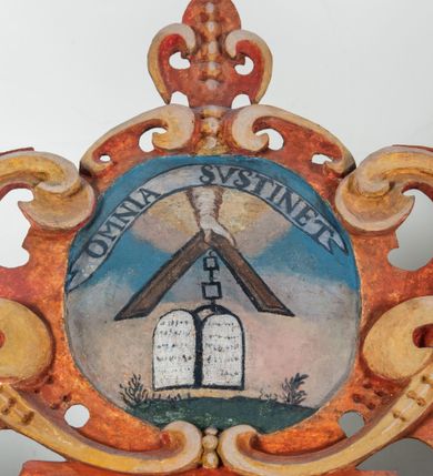 Zdjęcie nr 82: Umeblowanie zakrystii usytuowane jest wzdłuż ścian północnej, południowej i wschodniej pomieszczenia.  Na ścianie północnej, w części dolnej znajdują się trzy połączone ze sobą komody, każda o czterech szufladach, nakryte jednym blatem. W części górnej mieszczą się pięcioosiowe szafki z szufladami w cokole. Osie artykułowe są pilastrami hermowymi. Na osi środkowej szafka jest nieco wyższa i szersza z drzwiczkami dekorowanymi prostokątną, uszatą, oprofilowaną płyciną z malowanym przedstawieniem Chrystusa Eucharystycznego. Drzwiczki pozostałych szafek mają kształt stojących prostokątów i dekorowane są płycinami o wklęsłych narożach, zdobionymi gałązkami czerwonych róż, ułożonych w rokokowych wazonach. Ścianki zamknięte są niepełnym belkowaniem z gzymsem. W partii zwieńczenia osie artykułowane są cokolikami, na których ustawione są toczone, kuliste wazoniki. Pomiędzy nimi umieszczone są kartusze utworzone z ornamentu małżowinowo-chrząstkowego z malowanymi emblematami na owalnych medalionach, które ułożone są kolejno od lewej strony: 
1. Pionowo ustawiony miecz, ostrzem skierowany ku górze, połączony ze skrzyżowanymi berłem i dwoma kluczami, od których odchodzą fantazyjnie ułożone sznury w kolorze czerwonym. U góry napis na szarfie: „OBEDITE”.
2. W górnej części wyłaniająca się z chmur ręka trzymająca w dłoni węgielnicę i przymocowane do niej na łańcuszku tablice Dekalogu. Napis na szarfie: „OMNIA SVSTINET”.
3. Bóg Ojciec ukazany w półpostaci, lewą ręką opiera się na kuli ziemskiej, prawą czyni gest błogosławieństwa. 
4. Bujnie ulistnione, ciemnozielone drzewko spięte w dolnej części złotą formą w kształcie dwóch wywiniętych wolutek. Napis na szarfie: „GAVDET PATIENTIA DVRIS”. 
5. W górnej części obrazu z chmur wyłania się ręka, która zrzuca na ziemię iskrzący pocisk i dwa skrzyżowane pioruny, połączone parą białych skrzydeł. Napis „FORTIS VLTOR”.
 
Na ścianie środkowej, wschodniej znajduje się sarkofagowa mensa, powyżej parawanowa ścianka na profilowanym cokole, artykułowana pilastrami, pomiędzy którymi umieszczone są płyciny z kształcie leżących prostokątów z obrazami; ścianka zamknięta jest profilowanym gzymsem. W zwieńczeniu, na jej skrajach znajdują się owalne medaliony ujęte w kartusze z ornamentu małżowinowo-chrząstkowego. W medalionie po lewej stronie namalowane są litery alfa i omega na błękitnym tle oraz wstęga z napisem: „RENOVATUM A(NNO) D(OMINI) 1975”. Po prawej stronie analogiczny kartusz z literami w medalionie: „Ω / O / N” wpisane w formę krzyża, otoczonego niepełną aureolą; powyżej wstęga z napisem: „VIDENTEM AUDERE”. W płycinach obrazy przedstawiają świętych w ujęciu do pasa, zwróconych ku środkowi; od lewej:
1. Święty Demetriusz z włócznią opartą o prawy bark, ubrany w habit zakonu kanoników laterańskich od pokuty.
2. Święty Klet z aniołkiem, który trzyma w dłoni palmę męczeństwa i krzyż papieski. W prawym górnym narożu obrazu napis: „FAC HVIC SIGNO HONOREM”.
3. Święty Augustyn z gorejącym sercem na piersi i aniołkiem z łyżką i miseczką w dłoniach.
4. Święty ubrany w habit Marków, który trzyma w dłoni serce z krzyżem, podtrzymywane przez Chrystusa na obłokach – prawdopodobnie św. Cyriak, założyciel zakonu.
 
Na ścianie południowej znajduje się analogiczne umeblowanie, jak na ścianie północnej. W dolnej części dwie komody, a w miejscu trzeciej od wschodu szafka zdobiona dwiema uszatymi płycinami. Górna część mebla w przeciwieństwie do ściany północnej bez podwyższenia części środkowej. 
Na ścianie południowej:
1. Jasnobrązowa harfa na błękitnym tle i napis: „IN DOMINO”.
2. Ciemnobrązowy ptak i trzy podrywające się do lotu młode, w górnej części złote słońce i napis: „NON QUAE SVPER TERRAM.”
3. Po lewej stronie kompozycji wyłaniająca się z chmur ręka trzymająca skrzyżowane: miecz, czerwony krzyż i palmę. Napis: „NON ERVBESCO”.
4. W centrum przedstawienia krzak białej lilii o trzech rozgałęzieniach, w tle chmury w odcieniach koloru szaroniebieskiego. Napis na szarfie: „PER ARDVA DVLCIS”.
5. W lewej części kompozycji uskrzydlony jeleń, wspięty na tylnych nogach, po prawej stronie czarne i brązowe skały, w partii górnej otwarta korona, umieszczona pośrodku obłoku obwiedzionego biało-czerwonym pasem. Napis: „ET COMPREHENDATIS”.
 
W miejscu połączenia komód ścian północnej i południowej z dolną częścią konstrukcji na ścianie wschodniej znajdują się ukośnie ustawione, jednoskrzydłowe szafki o drzwiczkach dekorowanych prostokątną płyciną o wklęsłych narożach. Ścianki boczne części północnej i południowej zdobione są ukośną kratką. Powierzchnia szafek i komód pokryta jest polichromią imitującą marmur w kolorze beżowym, szarokremowym, różowym i rdzawym. Blaty malowane są farbą olejną w kolorze beżowym. Obramienia medalionów malowane w kolorze rdzawozłotym. Tło w większości medalionów w odcieniach błękitu, w górnych częściach majuskułowe napisy objaśniające przedstawienie, umieszczone w białych i półkoliście ułożonych szarfach. 