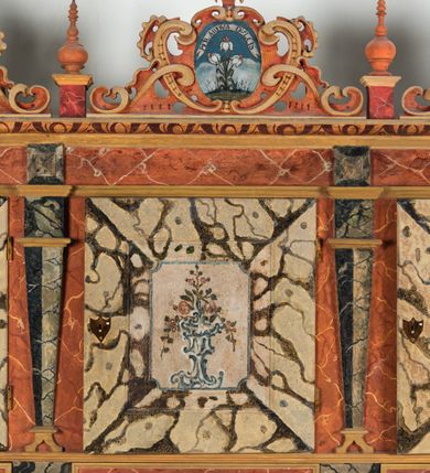 Zdjęcie nr 121: Umeblowanie zakrystii usytuowane jest wzdłuż ścian północnej, południowej i wschodniej pomieszczenia.  Na ścianie północnej, w części dolnej znajdują się trzy połączone ze sobą komody, każda o czterech szufladach, nakryte jednym blatem. W części górnej mieszczą się pięcioosiowe szafki z szufladami w cokole. Osie artykułowe są pilastrami hermowymi. Na osi środkowej szafka jest nieco wyższa i szersza z drzwiczkami dekorowanymi prostokątną, uszatą, oprofilowaną płyciną z malowanym przedstawieniem Chrystusa Eucharystycznego. Drzwiczki pozostałych szafek mają kształt stojących prostokątów i dekorowane są płycinami o wklęsłych narożach, zdobionymi gałązkami czerwonych róż, ułożonych w rokokowych wazonach. Ścianki zamknięte są niepełnym belkowaniem z gzymsem. W partii zwieńczenia osie artykułowane są cokolikami, na których ustawione są toczone, kuliste wazoniki. Pomiędzy nimi umieszczone są kartusze utworzone z ornamentu małżowinowo-chrząstkowego z malowanymi emblematami na owalnych medalionach, które ułożone są kolejno od lewej strony: 
1. Pionowo ustawiony miecz, ostrzem skierowany ku górze, połączony ze skrzyżowanymi berłem i dwoma kluczami, od których odchodzą fantazyjnie ułożone sznury w kolorze czerwonym. U góry napis na szarfie: „OBEDITE”.
2. W górnej części wyłaniająca się z chmur ręka trzymająca w dłoni węgielnicę i przymocowane do niej na łańcuszku tablice Dekalogu. Napis na szarfie: „OMNIA SVSTINET”.
3. Bóg Ojciec ukazany w półpostaci, lewą ręką opiera się na kuli ziemskiej, prawą czyni gest błogosławieństwa. 
4. Bujnie ulistnione, ciemnozielone drzewko spięte w dolnej części złotą formą w kształcie dwóch wywiniętych wolutek. Napis na szarfie: „GAVDET PATIENTIA DVRIS”. 
5. W górnej części obrazu z chmur wyłania się ręka, która zrzuca na ziemię iskrzący pocisk i dwa skrzyżowane pioruny, połączone parą białych skrzydeł. Napis „FORTIS VLTOR”.
 
Na ścianie środkowej, wschodniej znajduje się sarkofagowa mensa, powyżej parawanowa ścianka na profilowanym cokole, artykułowana pilastrami, pomiędzy którymi umieszczone są płyciny z kształcie leżących prostokątów z obrazami; ścianka zamknięta jest profilowanym gzymsem. W zwieńczeniu, na jej skrajach znajdują się owalne medaliony ujęte w kartusze z ornamentu małżowinowo-chrząstkowego. W medalionie po lewej stronie namalowane są litery alfa i omega na błękitnym tle oraz wstęga z napisem: „RENOVATUM A(NNO) D(OMINI) 1975”. Po prawej stronie analogiczny kartusz z literami w medalionie: „Ω / O / N” wpisane w formę krzyża, otoczonego niepełną aureolą; powyżej wstęga z napisem: „VIDENTEM AUDERE”. W płycinach obrazy przedstawiają świętych w ujęciu do pasa, zwróconych ku środkowi; od lewej:
1. Święty Demetriusz z włócznią opartą o prawy bark, ubrany w habit zakonu kanoników laterańskich od pokuty.
2. Święty Klet z aniołkiem, który trzyma w dłoni palmę męczeństwa i krzyż papieski. W prawym górnym narożu obrazu napis: „FAC HVIC SIGNO HONOREM”.
3. Święty Augustyn z gorejącym sercem na piersi i aniołkiem z łyżką i miseczką w dłoniach.
4. Święty ubrany w habit Marków, który trzyma w dłoni serce z krzyżem, podtrzymywane przez Chrystusa na obłokach – prawdopodobnie św. Cyriak, założyciel zakonu.
 
Na ścianie południowej znajduje się analogiczne umeblowanie, jak na ścianie północnej. W dolnej części dwie komody, a w miejscu trzeciej od wschodu szafka zdobiona dwiema uszatymi płycinami. Górna część mebla w przeciwieństwie do ściany północnej bez podwyższenia części środkowej. 
Na ścianie południowej:
1. Jasnobrązowa harfa na błękitnym tle i napis: „IN DOMINO”.
2. Ciemnobrązowy ptak i trzy podrywające się do lotu młode, w górnej części złote słońce i napis: „NON QUAE SVPER TERRAM.”
3. Po lewej stronie kompozycji wyłaniająca się z chmur ręka trzymająca skrzyżowane: miecz, czerwony krzyż i palmę. Napis: „NON ERVBESCO”.
4. W centrum przedstawienia krzak białej lilii o trzech rozgałęzieniach, w tle chmury w odcieniach koloru szaroniebieskiego. Napis na szarfie: „PER ARDVA DVLCIS”.
5. W lewej części kompozycji uskrzydlony jeleń, wspięty na tylnych nogach, po prawej stronie czarne i brązowe skały, w partii górnej otwarta korona, umieszczona pośrodku obłoku obwiedzionego biało-czerwonym pasem. Napis: „ET COMPREHENDATIS”.
 
W miejscu połączenia komód ścian północnej i południowej z dolną częścią konstrukcji na ścianie wschodniej znajdują się ukośnie ustawione, jednoskrzydłowe szafki o drzwiczkach dekorowanych prostokątną płyciną o wklęsłych narożach. Ścianki boczne części północnej i południowej zdobione są ukośną kratką. Powierzchnia szafek i komód pokryta jest polichromią imitującą marmur w kolorze beżowym, szarokremowym, różowym i rdzawym. Blaty malowane są farbą olejną w kolorze beżowym. Obramienia medalionów malowane w kolorze rdzawozłotym. Tło w większości medalionów w odcieniach błękitu, w górnych częściach majuskułowe napisy objaśniające przedstawienie, umieszczone w białych i półkoliście ułożonych szarfach. 