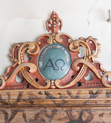 Zdjęcie nr 10: Umeblowanie zakrystii usytuowane jest wzdłuż ścian północnej, południowej i wschodniej pomieszczenia.  Na ścianie północnej, w części dolnej znajdują się trzy połączone ze sobą komody, każda o czterech szufladach, nakryte jednym blatem. W części górnej mieszczą się pięcioosiowe szafki z szufladami w cokole. Osie artykułowe są pilastrami hermowymi. Na osi środkowej szafka jest nieco wyższa i szersza z drzwiczkami dekorowanymi prostokątną, uszatą, oprofilowaną płyciną z malowanym przedstawieniem Chrystusa Eucharystycznego. Drzwiczki pozostałych szafek mają kształt stojących prostokątów i dekorowane są płycinami o wklęsłych narożach, zdobionymi gałązkami czerwonych róż, ułożonych w rokokowych wazonach. Ścianki zamknięte są niepełnym belkowaniem z gzymsem. W partii zwieńczenia osie artykułowane są cokolikami, na których ustawione są toczone, kuliste wazoniki. Pomiędzy nimi umieszczone są kartusze utworzone z ornamentu małżowinowo-chrząstkowego z malowanymi emblematami na owalnych medalionach, które ułożone są kolejno od lewej strony: 
1. Pionowo ustawiony miecz, ostrzem skierowany ku górze, połączony ze skrzyżowanymi berłem i dwoma kluczami, od których odchodzą fantazyjnie ułożone sznury w kolorze czerwonym. U góry napis na szarfie: „OBEDITE”.
2. W górnej części wyłaniająca się z chmur ręka trzymająca w dłoni węgielnicę i przymocowane do niej na łańcuszku tablice Dekalogu. Napis na szarfie: „OMNIA SVSTINET”.
3. Bóg Ojciec ukazany w półpostaci, lewą ręką opiera się na kuli ziemskiej, prawą czyni gest błogosławieństwa. 
4. Bujnie ulistnione, ciemnozielone drzewko spięte w dolnej części złotą formą w kształcie dwóch wywiniętych wolutek. Napis na szarfie: „GAVDET PATIENTIA DVRIS”. 
5. W górnej części obrazu z chmur wyłania się ręka, która zrzuca na ziemię iskrzący pocisk i dwa skrzyżowane pioruny, połączone parą białych skrzydeł. Napis „FORTIS VLTOR”.
 
Na ścianie środkowej, wschodniej znajduje się sarkofagowa mensa, powyżej parawanowa ścianka na profilowanym cokole, artykułowana pilastrami, pomiędzy którymi umieszczone są płyciny z kształcie leżących prostokątów z obrazami; ścianka zamknięta jest profilowanym gzymsem. W zwieńczeniu, na jej skrajach znajdują się owalne medaliony ujęte w kartusze z ornamentu małżowinowo-chrząstkowego. W medalionie po lewej stronie namalowane są litery alfa i omega na błękitnym tle oraz wstęga z napisem: „RENOVATUM A(NNO) D(OMINI) 1975”. Po prawej stronie analogiczny kartusz z literami w medalionie: „Ω / O / N” wpisane w formę krzyża, otoczonego niepełną aureolą; powyżej wstęga z napisem: „VIDENTEM AUDERE”. W płycinach obrazy przedstawiają świętych w ujęciu do pasa, zwróconych ku środkowi; od lewej:
1. Święty Demetriusz z włócznią opartą o prawy bark, ubrany w habit zakonu kanoników laterańskich od pokuty.
2. Święty Klet z aniołkiem, który trzyma w dłoni palmę męczeństwa i krzyż papieski. W prawym górnym narożu obrazu napis: „FAC HVIC SIGNO HONOREM”.
3. Święty Augustyn z gorejącym sercem na piersi i aniołkiem z łyżką i miseczką w dłoniach.
4. Święty ubrany w habit Marków, który trzyma w dłoni serce z krzyżem, podtrzymywane przez Chrystusa na obłokach – prawdopodobnie św. Cyriak, założyciel zakonu.
 
Na ścianie południowej znajduje się analogiczne umeblowanie, jak na ścianie północnej. W dolnej części dwie komody, a w miejscu trzeciej od wschodu szafka zdobiona dwiema uszatymi płycinami. Górna część mebla w przeciwieństwie do ściany północnej bez podwyższenia części środkowej. 
Na ścianie południowej:
1. Jasnobrązowa harfa na błękitnym tle i napis: „IN DOMINO”.
2. Ciemnobrązowy ptak i trzy podrywające się do lotu młode, w górnej części złote słońce i napis: „NON QUAE SVPER TERRAM.”
3. Po lewej stronie kompozycji wyłaniająca się z chmur ręka trzymająca skrzyżowane: miecz, czerwony krzyż i palmę. Napis: „NON ERVBESCO”.
4. W centrum przedstawienia krzak białej lilii o trzech rozgałęzieniach, w tle chmury w odcieniach koloru szaroniebieskiego. Napis na szarfie: „PER ARDVA DVLCIS”.
5. W lewej części kompozycji uskrzydlony jeleń, wspięty na tylnych nogach, po prawej stronie czarne i brązowe skały, w partii górnej otwarta korona, umieszczona pośrodku obłoku obwiedzionego biało-czerwonym pasem. Napis: „ET COMPREHENDATIS”.
 
W miejscu połączenia komód ścian północnej i południowej z dolną częścią konstrukcji na ścianie wschodniej znajdują się ukośnie ustawione, jednoskrzydłowe szafki o drzwiczkach dekorowanych prostokątną płyciną o wklęsłych narożach. Ścianki boczne części północnej i południowej zdobione są ukośną kratką. Powierzchnia szafek i komód pokryta jest polichromią imitującą marmur w kolorze beżowym, szarokremowym, różowym i rdzawym. Blaty malowane są farbą olejną w kolorze beżowym. Obramienia medalionów malowane w kolorze rdzawozłotym. Tło w większości medalionów w odcieniach błękitu, w górnych częściach majuskułowe napisy objaśniające przedstawienie, umieszczone w białych i półkoliście ułożonych szarfach. 