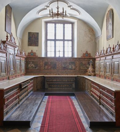 Zdjęcie nr 1: Umeblowanie zakrystii usytuowane jest wzdłuż ścian północnej, południowej i wschodniej pomieszczenia.  Na ścianie północnej, w części dolnej znajdują się trzy połączone ze sobą komody, każda o czterech szufladach, nakryte jednym blatem. W części górnej mieszczą się pięcioosiowe szafki z szufladami w cokole. Osie artykułowe są pilastrami hermowymi. Na osi środkowej szafka jest nieco wyższa i szersza z drzwiczkami dekorowanymi prostokątną, uszatą, oprofilowaną płyciną z malowanym przedstawieniem Chrystusa Eucharystycznego. Drzwiczki pozostałych szafek mają kształt stojących prostokątów i dekorowane są płycinami o wklęsłych narożach, zdobionymi gałązkami czerwonych róż, ułożonych w rokokowych wazonach. Ścianki zamknięte są niepełnym belkowaniem z gzymsem. W partii zwieńczenia osie artykułowane są cokolikami, na których ustawione są toczone, kuliste wazoniki. Pomiędzy nimi umieszczone są kartusze utworzone z ornamentu małżowinowo-chrząstkowego z malowanymi emblematami na owalnych medalionach, które ułożone są kolejno od lewej strony: 
1. Pionowo ustawiony miecz, ostrzem skierowany ku górze, połączony ze skrzyżowanymi berłem i dwoma kluczami, od których odchodzą fantazyjnie ułożone sznury w kolorze czerwonym. U góry napis na szarfie: „OBEDITE”.
2. W górnej części wyłaniająca się z chmur ręka trzymająca w dłoni węgielnicę i przymocowane do niej na łańcuszku tablice Dekalogu. Napis na szarfie: „OMNIA SVSTINET”.
3. Bóg Ojciec ukazany w półpostaci, lewą ręką opiera się na kuli ziemskiej, prawą czyni gest błogosławieństwa. 
4. Bujnie ulistnione, ciemnozielone drzewko spięte w dolnej części złotą formą w kształcie dwóch wywiniętych wolutek. Napis na szarfie: „GAVDET PATIENTIA DVRIS”. 
5. W górnej części obrazu z chmur wyłania się ręka, która zrzuca na ziemię iskrzący pocisk i dwa skrzyżowane pioruny, połączone parą białych skrzydeł. Napis „FORTIS VLTOR”.
 
Na ścianie środkowej, wschodniej znajduje się sarkofagowa mensa, powyżej parawanowa ścianka na profilowanym cokole, artykułowana pilastrami, pomiędzy którymi umieszczone są płyciny z kształcie leżących prostokątów z obrazami; ścianka zamknięta jest profilowanym gzymsem. W zwieńczeniu, na jej skrajach znajdują się owalne medaliony ujęte w kartusze z ornamentu małżowinowo-chrząstkowego. W medalionie po lewej stronie namalowane są litery alfa i omega na błękitnym tle oraz wstęga z napisem: „RENOVATUM A(NNO) D(OMINI) 1975”. Po prawej stronie analogiczny kartusz z literami w medalionie: „Ω / O / N” wpisane w formę krzyża, otoczonego niepełną aureolą; powyżej wstęga z napisem: „VIDENTEM AUDERE”. W płycinach obrazy przedstawiają świętych w ujęciu do pasa, zwróconych ku środkowi; od lewej:
1. Święty Demetriusz z włócznią opartą o prawy bark, ubrany w habit zakonu kanoników laterańskich od pokuty.
2. Święty Klet z aniołkiem, który trzyma w dłoni palmę męczeństwa i krzyż papieski. W prawym górnym narożu obrazu napis: „FAC HVIC SIGNO HONOREM”.
3. Święty Augustyn z gorejącym sercem na piersi i aniołkiem z łyżką i miseczką w dłoniach.
4. Święty ubrany w habit Marków, który trzyma w dłoni serce z krzyżem, podtrzymywane przez Chrystusa na obłokach – prawdopodobnie św. Cyriak, założyciel zakonu.
 
Na ścianie południowej znajduje się analogiczne umeblowanie, jak na ścianie północnej. W dolnej części dwie komody, a w miejscu trzeciej od wschodu szafka zdobiona dwiema uszatymi płycinami. Górna część mebla w przeciwieństwie do ściany północnej bez podwyższenia części środkowej. 
Na ścianie południowej:
1. Jasnobrązowa harfa na błękitnym tle i napis: „IN DOMINO”.
2. Ciemnobrązowy ptak i trzy podrywające się do lotu młode, w górnej części złote słońce i napis: „NON QUAE SVPER TERRAM.”
3. Po lewej stronie kompozycji wyłaniająca się z chmur ręka trzymająca skrzyżowane: miecz, czerwony krzyż i palmę. Napis: „NON ERVBESCO”.
4. W centrum przedstawienia krzak białej lilii o trzech rozgałęzieniach, w tle chmury w odcieniach koloru szaroniebieskiego. Napis na szarfie: „PER ARDVA DVLCIS”.
5. W lewej części kompozycji uskrzydlony jeleń, wspięty na tylnych nogach, po prawej stronie czarne i brązowe skały, w partii górnej otwarta korona, umieszczona pośrodku obłoku obwiedzionego biało-czerwonym pasem. Napis: „ET COMPREHENDATIS”.
 
W miejscu połączenia komód ścian północnej i południowej z dolną częścią konstrukcji na ścianie wschodniej znajdują się ukośnie ustawione, jednoskrzydłowe szafki o drzwiczkach dekorowanych prostokątną płyciną o wklęsłych narożach. Ścianki boczne części północnej i południowej zdobione są ukośną kratką. Powierzchnia szafek i komód pokryta jest polichromią imitującą marmur w kolorze beżowym, szarokremowym, różowym i rdzawym. Blaty malowane są farbą olejną w kolorze beżowym. Obramienia medalionów malowane w kolorze rdzawozłotym. Tło w większości medalionów w odcieniach błękitu, w górnych częściach majuskułowe napisy objaśniające przedstawienie, umieszczone w białych i półkoliście ułożonych szarfach. 