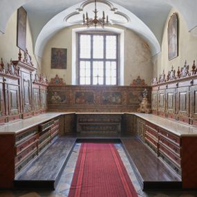 Zdjęcie nr 1: Umeblowanie zakrystii usytuowane jest wzdłuż ścian północnej, południowej i wschodniej pomieszczenia.  Na ścianie północnej, w części dolnej znajdują się trzy połączone ze sobą komody, każda o czterech szufladach, nakryte jednym blatem. W części górnej mieszczą się pięcioosiowe szafki z szufladami w cokole. Osie artykułowe są pilastrami hermowymi. Na osi środkowej szafka jest nieco wyższa i szersza z drzwiczkami dekorowanymi prostokątną, uszatą, oprofilowaną płyciną z malowanym przedstawieniem Chrystusa Eucharystycznego. Drzwiczki pozostałych szafek mają kształt stojących prostokątów i dekorowane są płycinami o wklęsłych narożach, zdobionymi gałązkami czerwonych róż, ułożonych w rokokowych wazonach. Ścianki zamknięte są niepełnym belkowaniem z gzymsem. W partii zwieńczenia osie artykułowane są cokolikami, na których ustawione są toczone, kuliste wazoniki. Pomiędzy nimi umieszczone są kartusze utworzone z ornamentu małżowinowo-chrząstkowego z malowanymi emblematami na owalnych medalionach, które ułożone są kolejno od lewej strony: 
1. Pionowo ustawiony miecz, ostrzem skierowany ku górze, połączony ze skrzyżowanymi berłem i dwoma kluczami, od których odchodzą fantazyjnie ułożone sznury w kolorze czerwonym. U góry napis na szarfie: „OBEDITE”.
2. W górnej części wyłaniająca się z chmur ręka trzymająca w dłoni węgielnicę i przymocowane do niej na łańcuszku tablice Dekalogu. Napis na szarfie: „OMNIA SVSTINET”.
3. Bóg Ojciec ukazany w półpostaci, lewą ręką opiera się na kuli ziemskiej, prawą czyni gest błogosławieństwa. 
4. Bujnie ulistnione, ciemnozielone drzewko spięte w dolnej części złotą formą w kształcie dwóch wywiniętych wolutek. Napis na szarfie: „GAVDET PATIENTIA DVRIS”. 
5. W górnej części obrazu z chmur wyłania się ręka, która zrzuca na ziemię iskrzący pocisk i dwa skrzyżowane pioruny, połączone parą białych skrzydeł. Napis „FORTIS VLTOR”.
 
Na ścianie środkowej, wschodniej znajduje się sarkofagowa mensa, powyżej parawanowa ścianka na profilowanym cokole, artykułowana pilastrami, pomiędzy którymi umieszczone są płyciny z kształcie leżących prostokątów z obrazami; ścianka zamknięta jest profilowanym gzymsem. W zwieńczeniu, na jej skrajach znajdują się owalne medaliony ujęte w kartusze z ornamentu małżowinowo-chrząstkowego. W medalionie po lewej stronie namalowane są litery alfa i omega na błękitnym tle oraz wstęga z napisem: „RENOVATUM A(NNO) D(OMINI) 1975”. Po prawej stronie analogiczny kartusz z literami w medalionie: „Ω / O / N” wpisane w formę krzyża, otoczonego niepełną aureolą; powyżej wstęga z napisem: „VIDENTEM AUDERE”. W płycinach obrazy przedstawiają świętych w ujęciu do pasa, zwróconych ku środkowi; od lewej:
1. Święty Demetriusz z włócznią opartą o prawy bark, ubrany w habit zakonu kanoników laterańskich od pokuty.
2. Święty Klet z aniołkiem, który trzyma w dłoni palmę męczeństwa i krzyż papieski. W prawym górnym narożu obrazu napis: „FAC HVIC SIGNO HONOREM”.
3. Święty Augustyn z gorejącym sercem na piersi i aniołkiem z łyżką i miseczką w dłoniach.
4. Święty ubrany w habit Marków, który trzyma w dłoni serce z krzyżem, podtrzymywane przez Chrystusa na obłokach – prawdopodobnie św. Cyriak, założyciel zakonu.
 
Na ścianie południowej znajduje się analogiczne umeblowanie, jak na ścianie północnej. W dolnej części dwie komody, a w miejscu trzeciej od wschodu szafka zdobiona dwiema uszatymi płycinami. Górna część mebla w przeciwieństwie do ściany północnej bez podwyższenia części środkowej. 
Na ścianie południowej:
1. Jasnobrązowa harfa na błękitnym tle i napis: „IN DOMINO”.
2. Ciemnobrązowy ptak i trzy podrywające się do lotu młode, w górnej części złote słońce i napis: „NON QUAE SVPER TERRAM.”
3. Po lewej stronie kompozycji wyłaniająca się z chmur ręka trzymająca skrzyżowane: miecz, czerwony krzyż i palmę. Napis: „NON ERVBESCO”.
4. W centrum przedstawienia krzak białej lilii o trzech rozgałęzieniach, w tle chmury w odcieniach koloru szaroniebieskiego. Napis na szarfie: „PER ARDVA DVLCIS”.
5. W lewej części kompozycji uskrzydlony jeleń, wspięty na tylnych nogach, po prawej stronie czarne i brązowe skały, w partii górnej otwarta korona, umieszczona pośrodku obłoku obwiedzionego biało-czerwonym pasem. Napis: „ET COMPREHENDATIS”.
 
W miejscu połączenia komód ścian północnej i południowej z dolną częścią konstrukcji na ścianie wschodniej znajdują się ukośnie ustawione, jednoskrzydłowe szafki o drzwiczkach dekorowanych prostokątną płyciną o wklęsłych narożach. Ścianki boczne części północnej i południowej zdobione są ukośną kratką. Powierzchnia szafek i komód pokryta jest polichromią imitującą marmur w kolorze beżowym, szarokremowym, różowym i rdzawym. Blaty malowane są farbą olejną w kolorze beżowym. Obramienia medalionów malowane w kolorze rdzawozłotym. Tło w większości medalionów w odcieniach błękitu, w górnych częściach majuskułowe napisy objaśniające przedstawienie, umieszczone w białych i półkoliście ułożonych szarfach. 