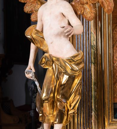 Zdjęcie nr 8: Rzeźba pełnoplastyczna, ścięta i drążona od tyłu.
Postać anioła o charakterystycznym wydłużeniu sylwetki, esowato wygięta i ustawiona na postumencie w lekkim kontrapoście;  ciężar ciała postaci wsparty na lewej nodze, lewa ręka zagięta w łokciu na wysokości pasa, prawa ręka podtrzymuje wydłużony kartusz. Nogi okryte są złotą draperią, która tworzy ekspresyjne, sztywne i lekko rozwiane fałdy. Opadająca z prawego ramienia szata obnaża goły tors i podbrzusze opadając na biodra. Drobna twarz skierowana jest na wprost, usta są pełne, lekko rozchylone, a pukle włosów opadają swobodnie na ramiona. Skrzydła są szeroko rozpostarte.