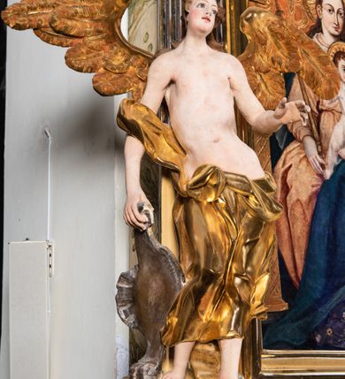 Zdjęcie nr 9: Rzeźba pełnoplastyczna, ścięta i drążona od tyłu.
Postać anioła o charakterystycznym wydłużeniu sylwetki, esowato wygięta i ustawiona na postumencie w lekkim kontrapoście;  ciężar ciała postaci wsparty na lewej nodze, lewa ręka zagięta w łokciu na wysokości pasa, prawa ręka podtrzymuje wydłużony kartusz. Nogi okryte są złotą draperią, która tworzy ekspresyjne, sztywne i lekko rozwiane fałdy. Opadająca z prawego ramienia szata obnaża goły tors i podbrzusze opadając na biodra. Drobna twarz skierowana jest na wprost, usta są pełne, lekko rozchylone, a pukle włosów opadają swobodnie na ramiona. Skrzydła są szeroko rozpostarte.