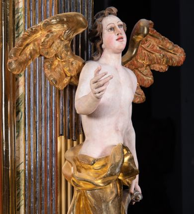 Zdjęcie nr 1: Rzeźba pełnoplastyczna, ścięta i drążona od tyłu.
Postać anioła o charakterystycznym wydłużeniu sylwetki, esowato wygięta i ustawiona na postumencie w lekkim kontrapoście;  ciężar ciała postaci wsparty na lewej nodze, lewa ręka zagięta w łokciu na wysokości pasa, prawa ręka podtrzymuje wydłużony kartusz. Nogi okryte są złotą draperią, która tworzy ekspresyjne, sztywne i lekko rozwiane fałdy. Opadająca z prawego ramienia szata obnaża goły tors i podbrzusze opadając na biodra. Drobna twarz skierowana jest na wprost, usta są pełne, lekko rozchylone, a pukle włosów opadają swobodnie na ramiona. Skrzydła są szeroko rozpostarte.