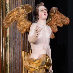 Zdjęcie nr 1: Rzeźba pełnoplastyczna, ścięta i drążona od tyłu.
Postać anioła o charakterystycznym wydłużeniu sylwetki, esowato wygięta i ustawiona na postumencie w lekkim kontrapoście;  ciężar ciała postaci wsparty na lewej nodze, lewa ręka zagięta w łokciu na wysokości pasa, prawa ręka podtrzymuje wydłużony kartusz. Nogi okryte są złotą draperią, która tworzy ekspresyjne, sztywne i lekko rozwiane fałdy. Opadająca z prawego ramienia szata obnaża goły tors i podbrzusze opadając na biodra. Drobna twarz skierowana jest na wprost, usta są pełne, lekko rozchylone, a pukle włosów opadają swobodnie na ramiona. Skrzydła są szeroko rozpostarte.