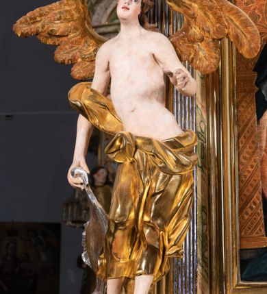 Zdjęcie nr 7: Rzeźba pełnoplastyczna, ścięta i drążona od tyłu.
Postać anioła o charakterystycznym wydłużeniu sylwetki, esowato wygięta i ustawiona na postumencie w lekkim kontrapoście;  ciężar ciała postaci wsparty na lewej nodze, lewa ręka zagięta w łokciu na wysokości pasa, prawa ręka podtrzymuje wydłużony kartusz. Nogi okryte są złotą draperią, która tworzy ekspresyjne, sztywne i lekko rozwiane fałdy. Opadająca z prawego ramienia szata obnaża goły tors i podbrzusze opadając na biodra. Drobna twarz skierowana jest na wprost, usta są pełne, lekko rozchylone, a pukle włosów opadają swobodnie na ramiona. Skrzydła są szeroko rozpostarte.