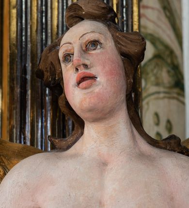 Zdjęcie nr 12: Rzeźba pełnoplastyczna, ścięta i drążona od tyłu.
Postać anioła o charakterystycznym wydłużeniu sylwetki, esowato wygięta i ustawiona na postumencie w lekkim kontrapoście;  ciężar ciała postaci wsparty na lewej nodze, lewa ręka zagięta w łokciu na wysokości pasa, prawa ręka podtrzymuje wydłużony kartusz. Nogi okryte są złotą draperią, która tworzy ekspresyjne, sztywne i lekko rozwiane fałdy. Opadająca z prawego ramienia szata obnaża goły tors i podbrzusze opadając na biodra. Drobna twarz skierowana jest na wprost, usta są pełne, lekko rozchylone, a pukle włosów opadają swobodnie na ramiona. Skrzydła są szeroko rozpostarte.