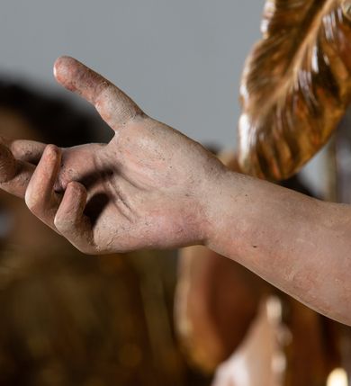 Zdjęcie nr 5: Rzeźba pełnoplastyczna, ścięta i drążona od tyłu.
Postać anioła o charakterystycznym wydłużeniu sylwetki, esowato wygięta i ustawiona na postumencie w lekkim kontrapoście;  ciężar ciała postaci wsparty na lewej nodze, lewa ręka zagięta w łokciu na wysokości pasa, prawa ręka podtrzymuje wydłużony kartusz. Nogi okryte są złotą draperią, która tworzy ekspresyjne, sztywne i lekko rozwiane fałdy. Opadająca z prawego ramienia szata obnaża goły tors i podbrzusze opadając na biodra. Drobna twarz skierowana jest na wprost, usta są pełne, lekko rozchylone, a pukle włosów opadają swobodnie na ramiona. Skrzydła są szeroko rozpostarte.