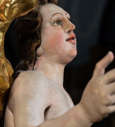 Zdjęcie nr 14: Rzeźba pełnoplastyczna, ścięta i drążona od tyłu.
Postać anioła o charakterystycznym wydłużeniu sylwetki, esowato wygięta i ustawiona na postumencie w lekkim kontrapoście;  ciężar ciała postaci wsparty na lewej nodze, lewa ręka zagięta w łokciu na wysokości pasa, prawa ręka podtrzymuje wydłużony kartusz. Nogi okryte są złotą draperią, która tworzy ekspresyjne, sztywne i lekko rozwiane fałdy. Opadająca z prawego ramienia szata obnaża goły tors i podbrzusze opadając na biodra. Drobna twarz skierowana jest na wprost, usta są pełne, lekko rozchylone, a pukle włosów opadają swobodnie na ramiona. Skrzydła są szeroko rozpostarte.