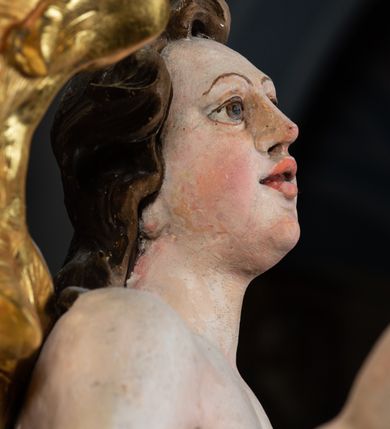 Zdjęcie nr 13: Rzeźba pełnoplastyczna, ścięta i drążona od tyłu.
Postać anioła o charakterystycznym wydłużeniu sylwetki, esowato wygięta i ustawiona na postumencie w lekkim kontrapoście;  ciężar ciała postaci wsparty na lewej nodze, lewa ręka zagięta w łokciu na wysokości pasa, prawa ręka podtrzymuje wydłużony kartusz. Nogi okryte są złotą draperią, która tworzy ekspresyjne, sztywne i lekko rozwiane fałdy. Opadająca z prawego ramienia szata obnaża goły tors i podbrzusze opadając na biodra. Drobna twarz skierowana jest na wprost, usta są pełne, lekko rozchylone, a pukle włosów opadają swobodnie na ramiona. Skrzydła są szeroko rozpostarte.