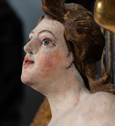 Zdjęcie nr 4: Rzeźba pełnoplastyczna, ścięta i drążona od tyłu.
Postać anioła o charakterystycznym wydłużeniu sylwetki, esowato wygięta i ustawiona na postumencie w lekkim kontrapoście;  ciężar ciała postaci wsparty na lewej nodze, lewa ręka zagięta w łokciu na wysokości pasa, prawa ręka podtrzymuje wydłużony kartusz. Nogi okryte są złotą draperią, która tworzy ekspresyjne, sztywne i lekko rozwiane fałdy. Opadająca z prawego ramienia szata obnaża goły tors i podbrzusze opadając na biodra. Drobna twarz skierowana jest na wprost, usta są pełne, lekko rozchylone, a pukle włosów opadają swobodnie na ramiona. Skrzydła są szeroko rozpostarte.