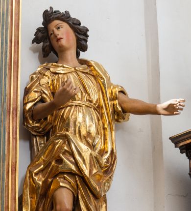 Zdjęcie nr 1: Rzeźba niemal pełnoplastyczna, ścięta. Stoi na konsoli małżowinowo chrząstkowej w lekkim kontrapoście z ciężarem ciała opartym na lewej nodze, ukazana jest frontalnie z wysuniętymi do przodu rękami ugiętymi w łokciach na wysokości pasa i głową przechyloną w prawo. Owalna, wydłużona twarz o pełnych policzkach, długim nosie, wąskich ustach i szeroko otwartych oczach, włosy krótkie faliste, rozwiane, zakrywające uszy. Rzeźba ubrana jest w tunikę z podwiniętymi rękawami, przepasaną na biodrze, odsłaniającą prawe udo i część lewej łydki oraz zarzucony na lewe ramię płaszcz z połą zarzuconą na prawe udo. Szaty modelowane ostrymi krawędziami, z podwiniętą dolną częścią płaszcza.