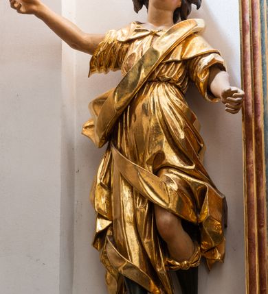 Zdjęcie nr 2: Forma niemal pełnoplastyczna i ścięta. Rzeźba stoi na konsoli małżowinowo chrząstkowej w lekkim kontrapoście z ciężarem ciała opartym na prawej nodze, ukazana jest frontalnie z wysuniętymi do przodu rękami, prawa uniesiona na wysokości głowy i znacznie odsunięta od tułowia, lewa lekko ugięta w łokciu na wysokości pasa.  Głowa przechylona w lewo. Okrągła twarz o pełnych policzkach i małym podbródku, długim nosie, wąskich, drobnych ustach i  szeroko otwartych oczach, włosy krótkie faliste, rozwiane, nieznacznie odkrywające uszy. Rzeźba ubrana jest w tunikę z podwiniętymi rękawami, przepasaną na biodrze, odsłaniającą lewe udo oraz w zarzucony na lewe ramię płaszcz z połą zarzuconą na prawe udo. Szaty modelowane ostrymi krawędziami, z podwiniętą dolną częścią płaszcza.