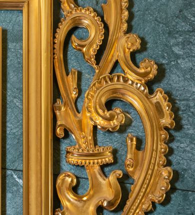 Zdjęcie nr 9: Obraz uzyskał kształt stojącego prostokąta ujętego w proste, złocone i profilowane ramy oraz dekoracyjne obramienie utworzone z płaskorzeźbionego ornamentu małżowinowo-chrząstkowego przytwierdzonego do zielono-błękitnego marmurowego tła. W zwieńczeniu obramienia kartusz z napisem: AVE MARIA. W centrum kompozycji na ciemnym, jednolitym tle ukazano frontalnie Madonnę z Dzieciątkiem w typie Matki Boskiej Częstochowskiej. Maria na lewym przedramieniu trzyma Dzieciątko Jezus, na które wskazuje prawą ręką przyłożoną do piersi. Matka Boska ma pociągłą twarz o brązowej karnacji z wyraźnie zaznaczonymi drobnymi ustami o pełnych wargach, wydatnym nosie i małych, migdałowych, otwartych oczach. Na jej prawym policzku widoczne są dwie rysy. Madonna ubrana jest w ciemnoniebieski maforion podbity czerwienią i lamowany taśmą z malarskim naśladownictwem kameryzacji. Spod maforionu widoczna jest zielona, marszczona suknia obwiedziona pod szyją taśmą także z malarskim naśladownictwem kameryzacji. Siedzące Dzieciątko Jezus zostało ujęte w trzech czwartych w prawo. Prawą rękę unosi w geście błogosławieństwa, lewą zaś podtrzymuje na kolanach zamknięty na dwie klamry kodeks. Jezus o jasnobrązowej karnacji ma owalną twarz o spojrzeniu skierowanym na matkę, mały nos i pełne usta. Jego głowę otaczają kędzierzawe, brązowe włosy rozjaśnione przy końcówkach loków. Ubrany jest w czerwoną, marszczoną na piersi tunikę lamowaną pod szyją, u spodu i przy rękawach taśmą z malarskim naśladownictwem kameryzacji. Spod tuniki wdać na dole jego bosą stopę. Głowy obu postaci otaczają złocone nimby oraz dwanaście malowanych gwiazd, na które nałożono mniejsze, metalowe i złocone. Na głowach Marii i Jezusa nałożone są srebrne i złocone korony o floranych sterczynach zdobione kameryzacją.    
