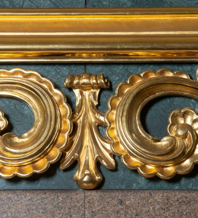 Zdjęcie nr 10: Obraz uzyskał kształt stojącego prostokąta ujętego w proste, złocone i profilowane ramy oraz dekoracyjne obramienie utworzone z płaskorzeźbionego ornamentu małżowinowo-chrząstkowego przytwierdzonego do zielono-błękitnego marmurowego tła. W zwieńczeniu obramienia kartusz z napisem: AVE MARIA. W centrum kompozycji na ciemnym, jednolitym tle ukazano frontalnie Madonnę z Dzieciątkiem w typie Matki Boskiej Częstochowskiej. Maria na lewym przedramieniu trzyma Dzieciątko Jezus, na które wskazuje prawą ręką przyłożoną do piersi. Matka Boska ma pociągłą twarz o brązowej karnacji z wyraźnie zaznaczonymi drobnymi ustami o pełnych wargach, wydatnym nosie i małych, migdałowych, otwartych oczach. Na jej prawym policzku widoczne są dwie rysy. Madonna ubrana jest w ciemnoniebieski maforion podbity czerwienią i lamowany taśmą z malarskim naśladownictwem kameryzacji. Spod maforionu widoczna jest zielona, marszczona suknia obwiedziona pod szyją taśmą także z malarskim naśladownictwem kameryzacji. Siedzące Dzieciątko Jezus zostało ujęte w trzech czwartych w prawo. Prawą rękę unosi w geście błogosławieństwa, lewą zaś podtrzymuje na kolanach zamknięty na dwie klamry kodeks. Jezus o jasnobrązowej karnacji ma owalną twarz o spojrzeniu skierowanym na matkę, mały nos i pełne usta. Jego głowę otaczają kędzierzawe, brązowe włosy rozjaśnione przy końcówkach loków. Ubrany jest w czerwoną, marszczoną na piersi tunikę lamowaną pod szyją, u spodu i przy rękawach taśmą z malarskim naśladownictwem kameryzacji. Spod tuniki wdać na dole jego bosą stopę. Głowy obu postaci otaczają złocone nimby oraz dwanaście malowanych gwiazd, na które nałożono mniejsze, metalowe i złocone. Na głowach Marii i Jezusa nałożone są srebrne i złocone korony o floranych sterczynach zdobione kameryzacją.    
