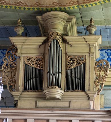 Zdjęcie nr 1: Chór muzyczny stanowi przyścienną, konstrukcję wspartą na dwóch jońskich kolumnach z przedpiersiem rozczłonkowanym balasami. Prospekt uzyskał formę trójosiowego prostopadłościanu flankowanego po bokach kolumnami (dziś niezachowanymi) i uszakami utworzonymi z ażurowej, złoconej wici suchego akantu. Na fazowanym cokole wyłamanym przy zewnętrznych krawędziach na wzór konsoli, usytuowano trójosiowe założenie zwieńczone profilowanym belkowaniem wyłamanym przy zewnętrznych krawędziach oraz przerwanym i podniesionym na osi środkowej. W osiach skrajnych w prostokątnych płycinach ustawiono piszczałki, które w górnej partii przykrywa ażurowy, złocony ornament z suchej wici akantowej. Na osi środkowej piszczałki wsparto na półkolistej, talerzowej konsoli, wieńcząc je półkolistym impostem wspierającym półkoliście artykułowane, zwielokrotnione belkowanie. W górnej części piszczałki zakrywa złocona, ażurowa wić utworzona z suchego akantu. Na belkowaniu bo bokach ustawiono wazy płomieniste. Całość prospektu utrzymana jest w jasnym kolorze.