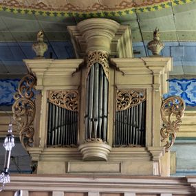 Zdjęcie nr 1: Chór muzyczny stanowi przyścienną, konstrukcję wspartą na dwóch jońskich kolumnach z przedpiersiem rozczłonkowanym balasami. Prospekt uzyskał formę trójosiowego prostopadłościanu flankowanego po bokach kolumnami (dziś niezachowanymi) i uszakami utworzonymi z ażurowej, złoconej wici suchego akantu. Na fazowanym cokole wyłamanym przy zewnętrznych krawędziach na wzór konsoli, usytuowano trójosiowe założenie zwieńczone profilowanym belkowaniem wyłamanym przy zewnętrznych krawędziach oraz przerwanym i podniesionym na osi środkowej. W osiach skrajnych w prostokątnych płycinach ustawiono piszczałki, które w górnej partii przykrywa ażurowy, złocony ornament z suchej wici akantowej. Na osi środkowej piszczałki wsparto na półkolistej, talerzowej konsoli, wieńcząc je półkolistym impostem wspierającym półkoliście artykułowane, zwielokrotnione belkowanie. W górnej części piszczałki zakrywa złocona, ażurowa wić utworzona z suchego akantu. Na belkowaniu bo bokach ustawiono wazy płomieniste. Całość prospektu utrzymana jest w jasnym kolorze.