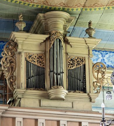 Zdjęcie nr 2: Chór muzyczny stanowi przyścienną, konstrukcję wspartą na dwóch jońskich kolumnach z przedpiersiem rozczłonkowanym balasami. Prospekt uzyskał formę trójosiowego prostopadłościanu flankowanego po bokach kolumnami (dziś niezachowanymi) i uszakami utworzonymi z ażurowej, złoconej wici suchego akantu. Na fazowanym cokole wyłamanym przy zewnętrznych krawędziach na wzór konsoli, usytuowano trójosiowe założenie zwieńczone profilowanym belkowaniem wyłamanym przy zewnętrznych krawędziach oraz przerwanym i podniesionym na osi środkowej. W osiach skrajnych w prostokątnych płycinach ustawiono piszczałki, które w górnej partii przykrywa ażurowy, złocony ornament z suchej wici akantowej. Na osi środkowej piszczałki wsparto na półkolistej, talerzowej konsoli, wieńcząc je półkolistym impostem wspierającym półkoliście artykułowane, zwielokrotnione belkowanie. W górnej części piszczałki zakrywa złocona, ażurowa wić utworzona z suchego akantu. Na belkowaniu bo bokach ustawiono wazy płomieniste. Całość prospektu utrzymana jest w jasnym kolorze.