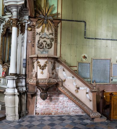 Zdjęcie nr 1: Przyścienną, wiszącą ambonę z dostępem w postaci jednobiegowych schodów z balustradą od strony prezbiterium usytuowano przy łuku tęczowym na ścianie północnej. Ambona składa się kosz na planie ściętego sześcioboku oraz zaplecka zwieńczonego gołębicą Ducha Świętego w promienistym otoku. Kosz ambony został w dolnej części zamknięty wyłamanym na jego ścięciach cokołem dekorowanym rytmicznie usytuowanymi płaskorzeźbionymi palmetami, tworząc dekoracyjny fryz, który poprowadzono również jako cokół balustrady schodów. Odpowiednikiem ukształtowania cokołu jest wieńczące kosz wyłamane belkowanie, także przechodzące na balustradę schodów, ozdobione w górnej partii mniejszymi liśćmi palmetowymi. Kosz od spodu zamknięty jest gładkimi spływami ujętymi wiszącą, liściastą szyszką. Ścięte boki kosza udekorowano spływami wolutowymi, ozdobionymi u spodu stylizowanym liściem akantu. W płycinach pomiędzy wolutami umieszczono złocone, wiszące gałązki różane. Zaplecek uzyskał kształt stojącego prostokąta flankowany dwoma pilastrami o głowicach pozbawionych dekoracji, na których wsparto przerwane belkowanie. W przerwie belkowania usytuowano złocony, promienisty otok z wyobrażeniem srebrzonej gołębicy Ducha Świętego. Trzony pilastrów zdobi wisząca gałązka liściasta, natomiast płycinę pomiędzy pilastrami motyw roślinny ułożony w formie arabeski. Strukturę pomalowano w dwóch odcieniach beżu, zaś elementy ornamentalne wyzłocono.