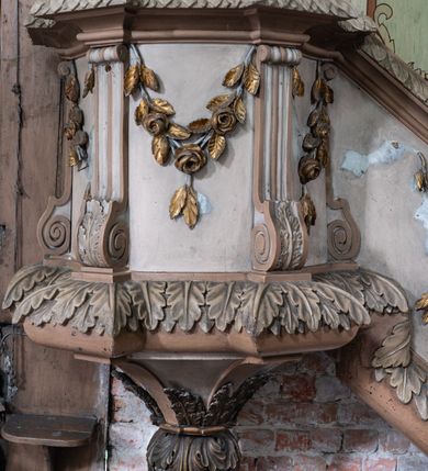 Zdjęcie nr 2: Przyścienną, wiszącą ambonę z dostępem w postaci jednobiegowych schodów z balustradą od strony prezbiterium usytuowano przy łuku tęczowym na ścianie północnej. Ambona składa się kosz na planie ściętego sześcioboku oraz zaplecka zwieńczonego gołębicą Ducha Świętego w promienistym otoku. Kosz ambony został w dolnej części zamknięty wyłamanym na jego ścięciach cokołem dekorowanym rytmicznie usytuowanymi płaskorzeźbionymi palmetami, tworząc dekoracyjny fryz, który poprowadzono również jako cokół balustrady schodów. Odpowiednikiem ukształtowania cokołu jest wieńczące kosz wyłamane belkowanie, także przechodzące na balustradę schodów, ozdobione w górnej partii mniejszymi liśćmi palmetowymi. Kosz od spodu zamknięty jest gładkimi spływami ujętymi wiszącą, liściastą szyszką. Ścięte boki kosza udekorowano spływami wolutowymi, ozdobionymi u spodu stylizowanym liściem akantu. W płycinach pomiędzy wolutami umieszczono złocone, wiszące gałązki różane. Zaplecek uzyskał kształt stojącego prostokąta flankowany dwoma pilastrami o głowicach pozbawionych dekoracji, na których wsparto przerwane belkowanie. W przerwie belkowania usytuowano złocony, promienisty otok z wyobrażeniem srebrzonej gołębicy Ducha Świętego. Trzony pilastrów zdobi wisząca gałązka liściasta, natomiast płycinę pomiędzy pilastrami motyw roślinny ułożony w formie arabeski. Strukturę pomalowano w dwóch odcieniach beżu, zaś elementy ornamentalne wyzłocono.