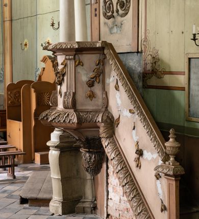 Zdjęcie nr 3: Przyścienną, wiszącą ambonę z dostępem w postaci jednobiegowych schodów z balustradą od strony prezbiterium usytuowano przy łuku tęczowym na ścianie północnej. Ambona składa się kosz na planie ściętego sześcioboku oraz zaplecka zwieńczonego gołębicą Ducha Świętego w promienistym otoku. Kosz ambony został w dolnej części zamknięty wyłamanym na jego ścięciach cokołem dekorowanym rytmicznie usytuowanymi płaskorzeźbionymi palmetami, tworząc dekoracyjny fryz, który poprowadzono również jako cokół balustrady schodów. Odpowiednikiem ukształtowania cokołu jest wieńczące kosz wyłamane belkowanie, także przechodzące na balustradę schodów, ozdobione w górnej partii mniejszymi liśćmi palmetowymi. Kosz od spodu zamknięty jest gładkimi spływami ujętymi wiszącą, liściastą szyszką. Ścięte boki kosza udekorowano spływami wolutowymi, ozdobionymi u spodu stylizowanym liściem akantu. W płycinach pomiędzy wolutami umieszczono złocone, wiszące gałązki różane. Zaplecek uzyskał kształt stojącego prostokąta flankowany dwoma pilastrami o głowicach pozbawionych dekoracji, na których wsparto przerwane belkowanie. W przerwie belkowania usytuowano złocony, promienisty otok z wyobrażeniem srebrzonej gołębicy Ducha Świętego. Trzony pilastrów zdobi wisząca gałązka liściasta, natomiast płycinę pomiędzy pilastrami motyw roślinny ułożony w formie arabeski. Strukturę pomalowano w dwóch odcieniach beżu, zaś elementy ornamentalne wyzłocono.