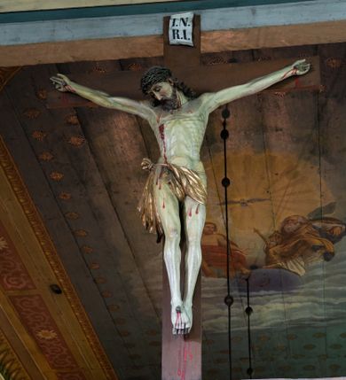 Zdjęcie nr 1: Rzeźbioną postać Ukrzyżowanego Chrystusa została umieszczona na drewnianym krzyżu, którego górne ramię wieńczy biały titulus w formie krótkiej banderoli z napisem: "I.N. / R.I". Ciało Chrystusa wisi na lekko opadniętych ramionach. Głowa Zbawiciela opada na jego prawe ramię. Chrystus ma pociągłą twarz okoloną kędzierzawą brodą oraz mocno pofalowanymi, ciemnymi włosami spływającymi na ramiona, odsłaniającymi jego lewe ucho. Na głowie ma koronę cierniową uplecioną z gęstych gałązek ciernia. Spod korony spływają na szyję i tułów strużki krwi. Anatomicznie ukształtowany tors wyraźnie ukazuje układ żeber oraz intensywna wybroczynę przebitego prawego boku. Nogi Zbawiciela, skrzyżowana jedna na drugiej, do krzyża zostały przytwierdzone jednym gwoździem, podobnie jak i obie dłonie. Biodra Jezusa okala złocone perizonium przerzucone przez sznur, z widocznym festonem przy prawym boku. Całość rzeźby pokryto naturalistyczną polichromią. 