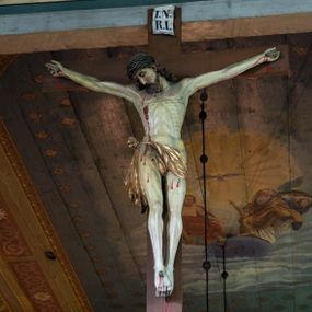 Zdjęcie nr 1: Rzeźbioną postać Ukrzyżowanego Chrystusa została umieszczona na drewnianym krzyżu, którego górne ramię wieńczy biały titulus w formie krótkiej banderoli z napisem: "I.N. / R.I". Ciało Chrystusa wisi na lekko opadniętych ramionach. Głowa Zbawiciela opada na jego prawe ramię. Chrystus ma pociągłą twarz okoloną kędzierzawą brodą oraz mocno pofalowanymi, ciemnymi włosami spływającymi na ramiona, odsłaniającymi jego lewe ucho. Na głowie ma koronę cierniową uplecioną z gęstych gałązek ciernia. Spod korony spływają na szyję i tułów strużki krwi. Anatomicznie ukształtowany tors wyraźnie ukazuje układ żeber oraz intensywna wybroczynę przebitego prawego boku. Nogi Zbawiciela, skrzyżowana jedna na drugiej, do krzyża zostały przytwierdzone jednym gwoździem, podobnie jak i obie dłonie. Biodra Jezusa okala złocone perizonium przerzucone przez sznur, z widocznym festonem przy prawym boku. Całość rzeźby pokryto naturalistyczną polichromią. 