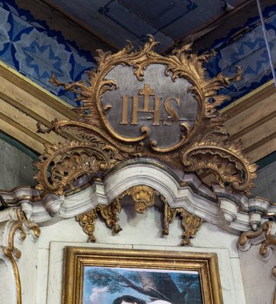 Zdjęcie nr 10: Przyścienny ołtarz Najświętszego Serca Pana Jezusa uzyskał formę architektonicznej nastawy jednokondygnacyjnej, jednoosiowej ze zwieńczeniem. Drewniana mensa z portatylem flankowana przez dwustrefowy wklęsło-wypukły cokół retabulum pierwotnie wspierała się na ażurowym, konsolowym stipesie ołtarzowym, dekorowanym ornamentami rocalliowymi. Kondygnacja wsparta na dwustrefowym cokole została ujęta dwiema parami pilastrów zwróconych delikatnie do wewnątrz oraz silnie wysuniętą do przodu parą kolumn o kapitelach kompozytowych. Podpory dźwigają belkowanie o wklęsło-wypukłej linii. Nad polem środkowym belkowanie jest przerwane, a na osiach podpór wyłamane. Na przedłużeniu kolumn na belkowaniu usytuowano wolutowe przyczółki dekorowane rocalliem, stanowiących postumenty do skradzionych figur aniołów. W polu środkowym umieszczono obraz Najświętszego Serca Pana Jezusa, ujęty profilowaną, złoconą ramą w formie stojącego prostokąta zamkniętego łukiem półkolistym nadwieszonym z uskokiem. Poniżej obrazu pomiędzy dwiema rocalliowymi konsolami widnieje zdjęcie obrazu św. Katarzyny Aleksandryjskiej. Zwieńczenie uzyskało formę leżącego prostokąta o wklęsłych, wolutowych bokach przechodzących u góry we wklęsło-wypukłe belkowanie, przyjmujące nad polem formę łuku falistego. W polu zwieńczenia znalazło się zdjęcie obrazu ze św. Sebastianem ujęte we wklęsło-wypukłą, profilowaną i złoconą ramę. Na szczycie w rocalliowym kartuszu wyobrażono złocony chrystogram "IHS" w srebrnym polu. Pilastry kondygnacji ozdobiono kapitelami z rocaille, a ich trzony udekorowano złoconymi zwisami utworzonymi z rocaille, rozet i liści. Całość struktury polichromowano na biało, a ornamenty i profile zostały wyzłocone. 