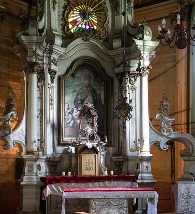 Zdjęcie nr 2: Kamienny ołtarz zamknięto w drewnianej obudowie, nakrywając go drewnianą mensą z portatylem. Stipes ołtarzowy uzyskał kształt prostopadłościanu. Antepedium ujęte zostało złoconą ramą dekorowaną rocaille’m. W jego polu umieszczono haftowaną tkaninę z medalionem w centrum z przedstawieniem w glorii chrystogramu "IHS", flankowany przez wić różaną. Na mensie ołtarza usytuowano tabernakulum w formie stojącego prostopadłościanu o płaskim froncie i wklęsłych bokach pozbawionych cokołu. Drzwiczki wprawiono w płaską niszę zwieńczoną profilowanym łukiem odcinkowym, nadwieszonym. U góry tabernakulum flankuje para sakramentek. Na tabernakulum usytuowano tron eucharystyczny utworzony z dwóch ażurowych spływów wolutowych podtrzymujących baldachim z lambrekinem. Na baldachimie na czerwonej poduszce spoczywa płaska, złocona korona otwarta. Retabulum ołtarzowe wzniesiono na planie półowalu i polichromowano je na biało. Jest to architektoniczna, jednokondygnacyjna, jednoosiowa struktura ze zwieńczeniem. Po bokach flankują ją bramki ustawione na dwustrefowym, tralkowym, wklęsło-wypukłym cokole. Łuki bramek tworzą fantazyjnie wygięte dwie woluty. Kondygnacja została ujęta dwiema parami pilastrów zwróconych delikatnie do wewnątrz oraz silnie wysuniętą do przodu parą kolumn o kapitelach kompozytowych. Podpory dźwigają pełne belkowanie o wklęsło-wypukłej linii. Nad polem środkowym belkowanie jest połączone gzymsem przyjmującym formę łuku falistego i wyłamane na osiach podpór. Kondygnację ujmują spływy wolutowe połączone z łukowato wygiętymi wolutami podtrzymywanymi przez wysokie tralkowe cokoły, tworząc bramki ołtarza. Bramki zwieńczono fantazyjnie wygiętymi impostami podtrzymującymi ornament rocalliowy. W polu środkowym umieszczono wielkoformatowe zdjęcie znajdującego się tu pierwotnie obrazu św. Mikołaja, ujęte profilowaną, złoconą ramą w formie stojącego prostokąta zamkniętego łukiem półkolistym nadwieszonym z uskokiem. Zwieńczenie uzyskało formę leżącego prostokąta flankowanego parą pilastrów zwróconych do wewnątrz oraz parą pilastrów zwróconych na zewnątrz, których trzony przechodzą w spływy wolutowe opadające na belkowanie. Pilastry dźwigają niepełne, wklęsło-wypukłe belkowanie przyjmujące nad polem formę łuku falistego. W polu zwieńczenia znalazło się przedstawienie gołębicy Ducha Świętego z ażurowymi promieniami na tle owalnego okna prezbiterium, ujęte w fantazjną, wklęsło-wypukłą, profilowaną i złoconą ramę. Pilastry kondygnacji ozdobiono kapitelami z rocaille, a ich trzony udekorowano złoconymi, rokokowymi panneau ze zwisami utworzonymi z rocaille, rozet i liści. Na ich tle usytuowano pierwotnie figury św. Kazimierza Królewicza i św. Augustyna (obecnie przeniesione do krypty nowego kościoła). Rzeźby stały na górnej strefie cokołu na wspornikach z rocaille. W przyłuczach i na belkowaniu znalazły się ornamenty roślinne. Także trzony pilastrów zwieńczenia dekorowane są złoconymi zwisami z rocaille i kwiatów. Pole wokół promieni z gołębicą Ducha Świętego również ozdobiono fragmentami rocaille. 