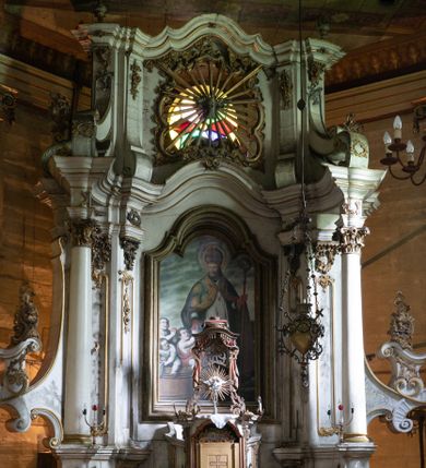 Zdjęcie nr 1: Kamienny ołtarz zamknięto w drewnianej obudowie, nakrywając go drewnianą mensą z portatylem. Stipes ołtarzowy uzyskał kształt prostopadłościanu. Antepedium ujęte zostało złoconą ramą dekorowaną rocaille’m. W jego polu umieszczono haftowaną tkaninę z medalionem w centrum z przedstawieniem w glorii chrystogramu &quot;IHS&quot;, flankowany przez wić różaną. Na mensie ołtarza usytuowano tabernakulum w formie stojącego prostopadłościanu o płaskim froncie i wklęsłych bokach pozbawionych cokołu. Drzwiczki wprawiono w płaską niszę zwieńczoną profilowanym łukiem odcinkowym, nadwieszonym. U góry tabernakulum flankuje para sakramentek. Na tabernakulum usytuowano tron eucharystyczny utworzony z dwóch ażurowych spływów wolutowych podtrzymujących baldachim z lambrekinem. Na baldachimie na czerwonej poduszce spoczywa płaska, złocona korona otwarta. Retabulum ołtarzowe wzniesiono na planie półowalu i polichromowano je na biało. Jest to architektoniczna, jednokondygnacyjna, jednoosiowa struktura ze zwieńczeniem. Po bokach flankują ją bramki ustawione na dwustrefowym, tralkowym, wklęsło-wypukłym cokole. Łuki bramek tworzą fantazyjnie wygięte dwie woluty. Kondygnacja została ujęta dwiema parami pilastrów zwróconych delikatnie do wewnątrz oraz silnie wysuniętą do przodu parą kolumn o kapitelach kompozytowych. Podpory dźwigają pełne belkowanie o wklęsło-wypukłej linii. Nad polem środkowym belkowanie jest połączone gzymsem przyjmującym formę łuku falistego i wyłamane na osiach podpór. Kondygnację ujmują spływy wolutowe połączone z łukowato wygiętymi wolutami podtrzymywanymi przez wysokie tralkowe cokoły, tworząc bramki ołtarza. Bramki zwieńczono fantazyjnie wygiętymi impostami podtrzymującymi ornament rocalliowy. W polu środkowym umieszczono wielkoformatowe zdjęcie znajdującego się tu pierwotnie obrazu św. Mikołaja, ujęte profilowaną, złoconą ramą w formie stojącego prostokąta zamkniętego łukiem półkolistym nadwieszonym z uskokiem. Zwieńczenie uzyskało formę leżącego prostokąta flankowanego parą pilastrów zwróconych do wewnątrz oraz parą pilastrów zwróconych na zewnątrz, których trzony przechodzą w spływy wolutowe opadające na belkowanie. Pilastry dźwigają niepełne, wklęsło-wypukłe belkowanie przyjmujące nad polem formę łuku falistego. W polu zwieńczenia znalazło się przedstawienie gołębicy Ducha Świętego z ażurowymi promieniami na tle owalnego okna prezbiterium, ujęte w fantazjną, wklęsło-wypukłą, profilowaną i złoconą ramę. Pilastry kondygnacji ozdobiono kapitelami z rocaille, a ich trzony udekorowano złoconymi, rokokowymi panneau ze zwisami utworzonymi z rocaille, rozet i liści. Na ich tle usytuowano pierwotnie figury św. Kazimierza Królewicza i św. Augustyna (obecnie przeniesione do krypty nowego kościoła). Rzeźby stały na górnej strefie cokołu na wspornikach z rocaille. W przyłuczach i na belkowaniu znalazły się ornamenty roślinne. Także trzony pilastrów zwieńczenia dekorowane są złoconymi zwisami z rocaille i kwiatów. Pole wokół promieni z gołębicą Ducha Świętego również ozdobiono fragmentami rocaille. 