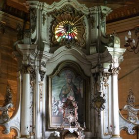 Zdjęcie nr 1: Kamienny ołtarz zamknięto w drewnianej obudowie, nakrywając go drewnianą mensą z portatylem. Stipes ołtarzowy uzyskał kształt prostopadłościanu. Antepedium ujęte zostało złoconą ramą dekorowaną rocaille’m. W jego polu umieszczono haftowaną tkaninę z medalionem w centrum z przedstawieniem w glorii chrystogramu "IHS", flankowany przez wić różaną. Na mensie ołtarza usytuowano tabernakulum w formie stojącego prostopadłościanu o płaskim froncie i wklęsłych bokach pozbawionych cokołu. Drzwiczki wprawiono w płaską niszę zwieńczoną profilowanym łukiem odcinkowym, nadwieszonym. U góry tabernakulum flankuje para sakramentek. Na tabernakulum usytuowano tron eucharystyczny utworzony z dwóch ażurowych spływów wolutowych podtrzymujących baldachim z lambrekinem. Na baldachimie na czerwonej poduszce spoczywa płaska, złocona korona otwarta. Retabulum ołtarzowe wzniesiono na planie półowalu i polichromowano je na biało. Jest to architektoniczna, jednokondygnacyjna, jednoosiowa struktura ze zwieńczeniem. Po bokach flankują ją bramki ustawione na dwustrefowym, tralkowym, wklęsło-wypukłym cokole. Łuki bramek tworzą fantazyjnie wygięte dwie woluty. Kondygnacja została ujęta dwiema parami pilastrów zwróconych delikatnie do wewnątrz oraz silnie wysuniętą do przodu parą kolumn o kapitelach kompozytowych. Podpory dźwigają pełne belkowanie o wklęsło-wypukłej linii. Nad polem środkowym belkowanie jest połączone gzymsem przyjmującym formę łuku falistego i wyłamane na osiach podpór. Kondygnację ujmują spływy wolutowe połączone z łukowato wygiętymi wolutami podtrzymywanymi przez wysokie tralkowe cokoły, tworząc bramki ołtarza. Bramki zwieńczono fantazyjnie wygiętymi impostami podtrzymującymi ornament rocalliowy. W polu środkowym umieszczono wielkoformatowe zdjęcie znajdującego się tu pierwotnie obrazu św. Mikołaja, ujęte profilowaną, złoconą ramą w formie stojącego prostokąta zamkniętego łukiem półkolistym nadwieszonym z uskokiem. Zwieńczenie uzyskało formę leżącego prostokąta flankowanego parą pilastrów zwróconych do wewnątrz oraz parą pilastrów zwróconych na zewnątrz, których trzony przechodzą w spływy wolutowe opadające na belkowanie. Pilastry dźwigają niepełne, wklęsło-wypukłe belkowanie przyjmujące nad polem formę łuku falistego. W polu zwieńczenia znalazło się przedstawienie gołębicy Ducha Świętego z ażurowymi promieniami na tle owalnego okna prezbiterium, ujęte w fantazjną, wklęsło-wypukłą, profilowaną i złoconą ramę. Pilastry kondygnacji ozdobiono kapitelami z rocaille, a ich trzony udekorowano złoconymi, rokokowymi panneau ze zwisami utworzonymi z rocaille, rozet i liści. Na ich tle usytuowano pierwotnie figury św. Kazimierza Królewicza i św. Augustyna (obecnie przeniesione do krypty nowego kościoła). Rzeźby stały na górnej strefie cokołu na wspornikach z rocaille. W przyłuczach i na belkowaniu znalazły się ornamenty roślinne. Także trzony pilastrów zwieńczenia dekorowane są złoconymi zwisami z rocaille i kwiatów. Pole wokół promieni z gołębicą Ducha Świętego również ozdobiono fragmentami rocaille. 