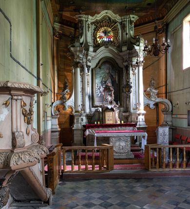 Zdjęcie nr 8: Kamienny ołtarz zamknięto w drewnianej obudowie, nakrywając go drewnianą mensą z portatylem. Stipes ołtarzowy uzyskał kształt prostopadłościanu. Antepedium ujęte zostało złoconą ramą dekorowaną rocaille’m. W jego polu umieszczono haftowaną tkaninę z medalionem w centrum z przedstawieniem w glorii chrystogramu &quot;IHS&quot;, flankowany przez wić różaną. Na mensie ołtarza usytuowano tabernakulum w formie stojącego prostopadłościanu o płaskim froncie i wklęsłych bokach pozbawionych cokołu. Drzwiczki wprawiono w płaską niszę zwieńczoną profilowanym łukiem odcinkowym, nadwieszonym. U góry tabernakulum flankuje para sakramentek. Na tabernakulum usytuowano tron eucharystyczny utworzony z dwóch ażurowych spływów wolutowych podtrzymujących baldachim z lambrekinem. Na baldachimie na czerwonej poduszce spoczywa płaska, złocona korona otwarta. Retabulum ołtarzowe wzniesiono na planie półowalu i polichromowano je na biało. Jest to architektoniczna, jednokondygnacyjna, jednoosiowa struktura ze zwieńczeniem. Po bokach flankują ją bramki ustawione na dwustrefowym, tralkowym, wklęsło-wypukłym cokole. Łuki bramek tworzą fantazyjnie wygięte dwie woluty. Kondygnacja została ujęta dwiema parami pilastrów zwróconych delikatnie do wewnątrz oraz silnie wysuniętą do przodu parą kolumn o kapitelach kompozytowych. Podpory dźwigają pełne belkowanie o wklęsło-wypukłej linii. Nad polem środkowym belkowanie jest połączone gzymsem przyjmującym formę łuku falistego i wyłamane na osiach podpór. Kondygnację ujmują spływy wolutowe połączone z łukowato wygiętymi wolutami podtrzymywanymi przez wysokie tralkowe cokoły, tworząc bramki ołtarza. Bramki zwieńczono fantazyjnie wygiętymi impostami podtrzymującymi ornament rocalliowy. W polu środkowym umieszczono wielkoformatowe zdjęcie znajdującego się tu pierwotnie obrazu św. Mikołaja, ujęte profilowaną, złoconą ramą w formie stojącego prostokąta zamkniętego łukiem półkolistym nadwieszonym z uskokiem. Zwieńczenie uzyskało formę leżącego prostokąta flankowanego parą pilastrów zwróconych do wewnątrz oraz parą pilastrów zwróconych na zewnątrz, których trzony przechodzą w spływy wolutowe opadające na belkowanie. Pilastry dźwigają niepełne, wklęsło-wypukłe belkowanie przyjmujące nad polem formę łuku falistego. W polu zwieńczenia znalazło się przedstawienie gołębicy Ducha Świętego z ażurowymi promieniami na tle owalnego okna prezbiterium, ujęte w fantazjną, wklęsło-wypukłą, profilowaną i złoconą ramę. Pilastry kondygnacji ozdobiono kapitelami z rocaille, a ich trzony udekorowano złoconymi, rokokowymi panneau ze zwisami utworzonymi z rocaille, rozet i liści. Na ich tle usytuowano pierwotnie figury św. Kazimierza Królewicza i św. Augustyna (obecnie przeniesione do krypty nowego kościoła). Rzeźby stały na górnej strefie cokołu na wspornikach z rocaille. W przyłuczach i na belkowaniu znalazły się ornamenty roślinne. Także trzony pilastrów zwieńczenia dekorowane są złoconymi zwisami z rocaille i kwiatów. Pole wokół promieni z gołębicą Ducha Świętego również ozdobiono fragmentami rocaille. 