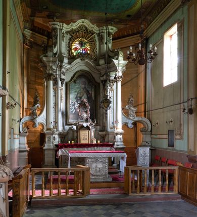 Zdjęcie nr 7: Kamienny ołtarz zamknięto w drewnianej obudowie, nakrywając go drewnianą mensą z portatylem. Stipes ołtarzowy uzyskał kształt prostopadłościanu. Antepedium ujęte zostało złoconą ramą dekorowaną rocaille’m. W jego polu umieszczono haftowaną tkaninę z medalionem w centrum z przedstawieniem w glorii chrystogramu &quot;IHS&quot;, flankowany przez wić różaną. Na mensie ołtarza usytuowano tabernakulum w formie stojącego prostopadłościanu o płaskim froncie i wklęsłych bokach pozbawionych cokołu. Drzwiczki wprawiono w płaską niszę zwieńczoną profilowanym łukiem odcinkowym, nadwieszonym. U góry tabernakulum flankuje para sakramentek. Na tabernakulum usytuowano tron eucharystyczny utworzony z dwóch ażurowych spływów wolutowych podtrzymujących baldachim z lambrekinem. Na baldachimie na czerwonej poduszce spoczywa płaska, złocona korona otwarta. Retabulum ołtarzowe wzniesiono na planie półowalu i polichromowano je na biało. Jest to architektoniczna, jednokondygnacyjna, jednoosiowa struktura ze zwieńczeniem. Po bokach flankują ją bramki ustawione na dwustrefowym, tralkowym, wklęsło-wypukłym cokole. Łuki bramek tworzą fantazyjnie wygięte dwie woluty. Kondygnacja została ujęta dwiema parami pilastrów zwróconych delikatnie do wewnątrz oraz silnie wysuniętą do przodu parą kolumn o kapitelach kompozytowych. Podpory dźwigają pełne belkowanie o wklęsło-wypukłej linii. Nad polem środkowym belkowanie jest połączone gzymsem przyjmującym formę łuku falistego i wyłamane na osiach podpór. Kondygnację ujmują spływy wolutowe połączone z łukowato wygiętymi wolutami podtrzymywanymi przez wysokie tralkowe cokoły, tworząc bramki ołtarza. Bramki zwieńczono fantazyjnie wygiętymi impostami podtrzymującymi ornament rocalliowy. W polu środkowym umieszczono wielkoformatowe zdjęcie znajdującego się tu pierwotnie obrazu św. Mikołaja, ujęte profilowaną, złoconą ramą w formie stojącego prostokąta zamkniętego łukiem półkolistym nadwieszonym z uskokiem. Zwieńczenie uzyskało formę leżącego prostokąta flankowanego parą pilastrów zwróconych do wewnątrz oraz parą pilastrów zwróconych na zewnątrz, których trzony przechodzą w spływy wolutowe opadające na belkowanie. Pilastry dźwigają niepełne, wklęsło-wypukłe belkowanie przyjmujące nad polem formę łuku falistego. W polu zwieńczenia znalazło się przedstawienie gołębicy Ducha Świętego z ażurowymi promieniami na tle owalnego okna prezbiterium, ujęte w fantazjną, wklęsło-wypukłą, profilowaną i złoconą ramę. Pilastry kondygnacji ozdobiono kapitelami z rocaille, a ich trzony udekorowano złoconymi, rokokowymi panneau ze zwisami utworzonymi z rocaille, rozet i liści. Na ich tle usytuowano pierwotnie figury św. Kazimierza Królewicza i św. Augustyna (obecnie przeniesione do krypty nowego kościoła). Rzeźby stały na górnej strefie cokołu na wspornikach z rocaille. W przyłuczach i na belkowaniu znalazły się ornamenty roślinne. Także trzony pilastrów zwieńczenia dekorowane są złoconymi zwisami z rocaille i kwiatów. Pole wokół promieni z gołębicą Ducha Świętego również ozdobiono fragmentami rocaille. 