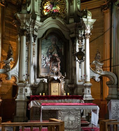 Zdjęcie nr 9: Kamienny ołtarz zamknięto w drewnianej obudowie, nakrywając go drewnianą mensą z portatylem. Stipes ołtarzowy uzyskał kształt prostopadłościanu. Antepedium ujęte zostało złoconą ramą dekorowaną rocaille’m. W jego polu umieszczono haftowaną tkaninę z medalionem w centrum z przedstawieniem w glorii chrystogramu "IHS", flankowany przez wić różaną. Na mensie ołtarza usytuowano tabernakulum w formie stojącego prostopadłościanu o płaskim froncie i wklęsłych bokach pozbawionych cokołu. Drzwiczki wprawiono w płaską niszę zwieńczoną profilowanym łukiem odcinkowym, nadwieszonym. U góry tabernakulum flankuje para sakramentek. Na tabernakulum usytuowano tron eucharystyczny utworzony z dwóch ażurowych spływów wolutowych podtrzymujących baldachim z lambrekinem. Na baldachimie na czerwonej poduszce spoczywa płaska, złocona korona otwarta. Retabulum ołtarzowe wzniesiono na planie półowalu i polichromowano je na biało. Jest to architektoniczna, jednokondygnacyjna, jednoosiowa struktura ze zwieńczeniem. Po bokach flankują ją bramki ustawione na dwustrefowym, tralkowym, wklęsło-wypukłym cokole. Łuki bramek tworzą fantazyjnie wygięte dwie woluty. Kondygnacja została ujęta dwiema parami pilastrów zwróconych delikatnie do wewnątrz oraz silnie wysuniętą do przodu parą kolumn o kapitelach kompozytowych. Podpory dźwigają pełne belkowanie o wklęsło-wypukłej linii. Nad polem środkowym belkowanie jest połączone gzymsem przyjmującym formę łuku falistego i wyłamane na osiach podpór. Kondygnację ujmują spływy wolutowe połączone z łukowato wygiętymi wolutami podtrzymywanymi przez wysokie tralkowe cokoły, tworząc bramki ołtarza. Bramki zwieńczono fantazyjnie wygiętymi impostami podtrzymującymi ornament rocalliowy. W polu środkowym umieszczono wielkoformatowe zdjęcie znajdującego się tu pierwotnie obrazu św. Mikołaja, ujęte profilowaną, złoconą ramą w formie stojącego prostokąta zamkniętego łukiem półkolistym nadwieszonym z uskokiem. Zwieńczenie uzyskało formę leżącego prostokąta flankowanego parą pilastrów zwróconych do wewnątrz oraz parą pilastrów zwróconych na zewnątrz, których trzony przechodzą w spływy wolutowe opadające na belkowanie. Pilastry dźwigają niepełne, wklęsło-wypukłe belkowanie przyjmujące nad polem formę łuku falistego. W polu zwieńczenia znalazło się przedstawienie gołębicy Ducha Świętego z ażurowymi promieniami na tle owalnego okna prezbiterium, ujęte w fantazjną, wklęsło-wypukłą, profilowaną i złoconą ramę. Pilastry kondygnacji ozdobiono kapitelami z rocaille, a ich trzony udekorowano złoconymi, rokokowymi panneau ze zwisami utworzonymi z rocaille, rozet i liści. Na ich tle usytuowano pierwotnie figury św. Kazimierza Królewicza i św. Augustyna (obecnie przeniesione do krypty nowego kościoła). Rzeźby stały na górnej strefie cokołu na wspornikach z rocaille. W przyłuczach i na belkowaniu znalazły się ornamenty roślinne. Także trzony pilastrów zwieńczenia dekorowane są złoconymi zwisami z rocaille i kwiatów. Pole wokół promieni z gołębicą Ducha Świętego również ozdobiono fragmentami rocaille. 