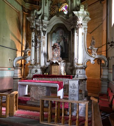 Zdjęcie nr 10: Kamienny ołtarz zamknięto w drewnianej obudowie, nakrywając go drewnianą mensą z portatylem. Stipes ołtarzowy uzyskał kształt prostopadłościanu. Antepedium ujęte zostało złoconą ramą dekorowaną rocaille’m. W jego polu umieszczono haftowaną tkaninę z medalionem w centrum z przedstawieniem w glorii chrystogramu &quot;IHS&quot;, flankowany przez wić różaną. Na mensie ołtarza usytuowano tabernakulum w formie stojącego prostopadłościanu o płaskim froncie i wklęsłych bokach pozbawionych cokołu. Drzwiczki wprawiono w płaską niszę zwieńczoną profilowanym łukiem odcinkowym, nadwieszonym. U góry tabernakulum flankuje para sakramentek. Na tabernakulum usytuowano tron eucharystyczny utworzony z dwóch ażurowych spływów wolutowych podtrzymujących baldachim z lambrekinem. Na baldachimie na czerwonej poduszce spoczywa płaska, złocona korona otwarta. Retabulum ołtarzowe wzniesiono na planie półowalu i polichromowano je na biało. Jest to architektoniczna, jednokondygnacyjna, jednoosiowa struktura ze zwieńczeniem. Po bokach flankują ją bramki ustawione na dwustrefowym, tralkowym, wklęsło-wypukłym cokole. Łuki bramek tworzą fantazyjnie wygięte dwie woluty. Kondygnacja została ujęta dwiema parami pilastrów zwróconych delikatnie do wewnątrz oraz silnie wysuniętą do przodu parą kolumn o kapitelach kompozytowych. Podpory dźwigają pełne belkowanie o wklęsło-wypukłej linii. Nad polem środkowym belkowanie jest połączone gzymsem przyjmującym formę łuku falistego i wyłamane na osiach podpór. Kondygnację ujmują spływy wolutowe połączone z łukowato wygiętymi wolutami podtrzymywanymi przez wysokie tralkowe cokoły, tworząc bramki ołtarza. Bramki zwieńczono fantazyjnie wygiętymi impostami podtrzymującymi ornament rocalliowy. W polu środkowym umieszczono wielkoformatowe zdjęcie znajdującego się tu pierwotnie obrazu św. Mikołaja, ujęte profilowaną, złoconą ramą w formie stojącego prostokąta zamkniętego łukiem półkolistym nadwieszonym z uskokiem. Zwieńczenie uzyskało formę leżącego prostokąta flankowanego parą pilastrów zwróconych do wewnątrz oraz parą pilastrów zwróconych na zewnątrz, których trzony przechodzą w spływy wolutowe opadające na belkowanie. Pilastry dźwigają niepełne, wklęsło-wypukłe belkowanie przyjmujące nad polem formę łuku falistego. W polu zwieńczenia znalazło się przedstawienie gołębicy Ducha Świętego z ażurowymi promieniami na tle owalnego okna prezbiterium, ujęte w fantazjną, wklęsło-wypukłą, profilowaną i złoconą ramę. Pilastry kondygnacji ozdobiono kapitelami z rocaille, a ich trzony udekorowano złoconymi, rokokowymi panneau ze zwisami utworzonymi z rocaille, rozet i liści. Na ich tle usytuowano pierwotnie figury św. Kazimierza Królewicza i św. Augustyna (obecnie przeniesione do krypty nowego kościoła). Rzeźby stały na górnej strefie cokołu na wspornikach z rocaille. W przyłuczach i na belkowaniu znalazły się ornamenty roślinne. Także trzony pilastrów zwieńczenia dekorowane są złoconymi zwisami z rocaille i kwiatów. Pole wokół promieni z gołębicą Ducha Świętego również ozdobiono fragmentami rocaille. 