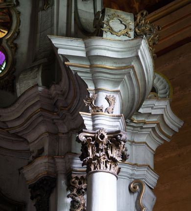 Zdjęcie nr 12: Kamienny ołtarz zamknięto w drewnianej obudowie, nakrywając go drewnianą mensą z portatylem. Stipes ołtarzowy uzyskał kształt prostopadłościanu. Antepedium ujęte zostało złoconą ramą dekorowaną rocaille’m. W jego polu umieszczono haftowaną tkaninę z medalionem w centrum z przedstawieniem w glorii chrystogramu &quot;IHS&quot;, flankowany przez wić różaną. Na mensie ołtarza usytuowano tabernakulum w formie stojącego prostopadłościanu o płaskim froncie i wklęsłych bokach pozbawionych cokołu. Drzwiczki wprawiono w płaską niszę zwieńczoną profilowanym łukiem odcinkowym, nadwieszonym. U góry tabernakulum flankuje para sakramentek. Na tabernakulum usytuowano tron eucharystyczny utworzony z dwóch ażurowych spływów wolutowych podtrzymujących baldachim z lambrekinem. Na baldachimie na czerwonej poduszce spoczywa płaska, złocona korona otwarta. Retabulum ołtarzowe wzniesiono na planie półowalu i polichromowano je na biało. Jest to architektoniczna, jednokondygnacyjna, jednoosiowa struktura ze zwieńczeniem. Po bokach flankują ją bramki ustawione na dwustrefowym, tralkowym, wklęsło-wypukłym cokole. Łuki bramek tworzą fantazyjnie wygięte dwie woluty. Kondygnacja została ujęta dwiema parami pilastrów zwróconych delikatnie do wewnątrz oraz silnie wysuniętą do przodu parą kolumn o kapitelach kompozytowych. Podpory dźwigają pełne belkowanie o wklęsło-wypukłej linii. Nad polem środkowym belkowanie jest połączone gzymsem przyjmującym formę łuku falistego i wyłamane na osiach podpór. Kondygnację ujmują spływy wolutowe połączone z łukowato wygiętymi wolutami podtrzymywanymi przez wysokie tralkowe cokoły, tworząc bramki ołtarza. Bramki zwieńczono fantazyjnie wygiętymi impostami podtrzymującymi ornament rocalliowy. W polu środkowym umieszczono wielkoformatowe zdjęcie znajdującego się tu pierwotnie obrazu św. Mikołaja, ujęte profilowaną, złoconą ramą w formie stojącego prostokąta zamkniętego łukiem półkolistym nadwieszonym z uskokiem. Zwieńczenie uzyskało formę leżącego prostokąta flankowanego parą pilastrów zwróconych do wewnątrz oraz parą pilastrów zwróconych na zewnątrz, których trzony przechodzą w spływy wolutowe opadające na belkowanie. Pilastry dźwigają niepełne, wklęsło-wypukłe belkowanie przyjmujące nad polem formę łuku falistego. W polu zwieńczenia znalazło się przedstawienie gołębicy Ducha Świętego z ażurowymi promieniami na tle owalnego okna prezbiterium, ujęte w fantazjną, wklęsło-wypukłą, profilowaną i złoconą ramę. Pilastry kondygnacji ozdobiono kapitelami z rocaille, a ich trzony udekorowano złoconymi, rokokowymi panneau ze zwisami utworzonymi z rocaille, rozet i liści. Na ich tle usytuowano pierwotnie figury św. Kazimierza Królewicza i św. Augustyna (obecnie przeniesione do krypty nowego kościoła). Rzeźby stały na górnej strefie cokołu na wspornikach z rocaille. W przyłuczach i na belkowaniu znalazły się ornamenty roślinne. Także trzony pilastrów zwieńczenia dekorowane są złoconymi zwisami z rocaille i kwiatów. Pole wokół promieni z gołębicą Ducha Świętego również ozdobiono fragmentami rocaille. 