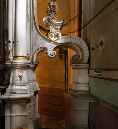 Zdjęcie nr 5: Kamienny ołtarz zamknięto w drewnianej obudowie, nakrywając go drewnianą mensą z portatylem. Stipes ołtarzowy uzyskał kształt prostopadłościanu. Antepedium ujęte zostało złoconą ramą dekorowaną rocaille’m. W jego polu umieszczono haftowaną tkaninę z medalionem w centrum z przedstawieniem w glorii chrystogramu "IHS", flankowany przez wić różaną. Na mensie ołtarza usytuowano tabernakulum w formie stojącego prostopadłościanu o płaskim froncie i wklęsłych bokach pozbawionych cokołu. Drzwiczki wprawiono w płaską niszę zwieńczoną profilowanym łukiem odcinkowym, nadwieszonym. U góry tabernakulum flankuje para sakramentek. Na tabernakulum usytuowano tron eucharystyczny utworzony z dwóch ażurowych spływów wolutowych podtrzymujących baldachim z lambrekinem. Na baldachimie na czerwonej poduszce spoczywa płaska, złocona korona otwarta. Retabulum ołtarzowe wzniesiono na planie półowalu i polichromowano je na biało. Jest to architektoniczna, jednokondygnacyjna, jednoosiowa struktura ze zwieńczeniem. Po bokach flankują ją bramki ustawione na dwustrefowym, tralkowym, wklęsło-wypukłym cokole. Łuki bramek tworzą fantazyjnie wygięte dwie woluty. Kondygnacja została ujęta dwiema parami pilastrów zwróconych delikatnie do wewnątrz oraz silnie wysuniętą do przodu parą kolumn o kapitelach kompozytowych. Podpory dźwigają pełne belkowanie o wklęsło-wypukłej linii. Nad polem środkowym belkowanie jest połączone gzymsem przyjmującym formę łuku falistego i wyłamane na osiach podpór. Kondygnację ujmują spływy wolutowe połączone z łukowato wygiętymi wolutami podtrzymywanymi przez wysokie tralkowe cokoły, tworząc bramki ołtarza. Bramki zwieńczono fantazyjnie wygiętymi impostami podtrzymującymi ornament rocalliowy. W polu środkowym umieszczono wielkoformatowe zdjęcie znajdującego się tu pierwotnie obrazu św. Mikołaja, ujęte profilowaną, złoconą ramą w formie stojącego prostokąta zamkniętego łukiem półkolistym nadwieszonym z uskokiem. Zwieńczenie uzyskało formę leżącego prostokąta flankowanego parą pilastrów zwróconych do wewnątrz oraz parą pilastrów zwróconych na zewnątrz, których trzony przechodzą w spływy wolutowe opadające na belkowanie. Pilastry dźwigają niepełne, wklęsło-wypukłe belkowanie przyjmujące nad polem formę łuku falistego. W polu zwieńczenia znalazło się przedstawienie gołębicy Ducha Świętego z ażurowymi promieniami na tle owalnego okna prezbiterium, ujęte w fantazjną, wklęsło-wypukłą, profilowaną i złoconą ramę. Pilastry kondygnacji ozdobiono kapitelami z rocaille, a ich trzony udekorowano złoconymi, rokokowymi panneau ze zwisami utworzonymi z rocaille, rozet i liści. Na ich tle usytuowano pierwotnie figury św. Kazimierza Królewicza i św. Augustyna (obecnie przeniesione do krypty nowego kościoła). Rzeźby stały na górnej strefie cokołu na wspornikach z rocaille. W przyłuczach i na belkowaniu znalazły się ornamenty roślinne. Także trzony pilastrów zwieńczenia dekorowane są złoconymi zwisami z rocaille i kwiatów. Pole wokół promieni z gołębicą Ducha Świętego również ozdobiono fragmentami rocaille. 