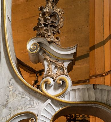 Zdjęcie nr 6: Kamienny ołtarz zamknięto w drewnianej obudowie, nakrywając go drewnianą mensą z portatylem. Stipes ołtarzowy uzyskał kształt prostopadłościanu. Antepedium ujęte zostało złoconą ramą dekorowaną rocaille’m. W jego polu umieszczono haftowaną tkaninę z medalionem w centrum z przedstawieniem w glorii chrystogramu &quot;IHS&quot;, flankowany przez wić różaną. Na mensie ołtarza usytuowano tabernakulum w formie stojącego prostopadłościanu o płaskim froncie i wklęsłych bokach pozbawionych cokołu. Drzwiczki wprawiono w płaską niszę zwieńczoną profilowanym łukiem odcinkowym, nadwieszonym. U góry tabernakulum flankuje para sakramentek. Na tabernakulum usytuowano tron eucharystyczny utworzony z dwóch ażurowych spływów wolutowych podtrzymujących baldachim z lambrekinem. Na baldachimie na czerwonej poduszce spoczywa płaska, złocona korona otwarta. Retabulum ołtarzowe wzniesiono na planie półowalu i polichromowano je na biało. Jest to architektoniczna, jednokondygnacyjna, jednoosiowa struktura ze zwieńczeniem. Po bokach flankują ją bramki ustawione na dwustrefowym, tralkowym, wklęsło-wypukłym cokole. Łuki bramek tworzą fantazyjnie wygięte dwie woluty. Kondygnacja została ujęta dwiema parami pilastrów zwróconych delikatnie do wewnątrz oraz silnie wysuniętą do przodu parą kolumn o kapitelach kompozytowych. Podpory dźwigają pełne belkowanie o wklęsło-wypukłej linii. Nad polem środkowym belkowanie jest połączone gzymsem przyjmującym formę łuku falistego i wyłamane na osiach podpór. Kondygnację ujmują spływy wolutowe połączone z łukowato wygiętymi wolutami podtrzymywanymi przez wysokie tralkowe cokoły, tworząc bramki ołtarza. Bramki zwieńczono fantazyjnie wygiętymi impostami podtrzymującymi ornament rocalliowy. W polu środkowym umieszczono wielkoformatowe zdjęcie znajdującego się tu pierwotnie obrazu św. Mikołaja, ujęte profilowaną, złoconą ramą w formie stojącego prostokąta zamkniętego łukiem półkolistym nadwieszonym z uskokiem. Zwieńczenie uzyskało formę leżącego prostokąta flankowanego parą pilastrów zwróconych do wewnątrz oraz parą pilastrów zwróconych na zewnątrz, których trzony przechodzą w spływy wolutowe opadające na belkowanie. Pilastry dźwigają niepełne, wklęsło-wypukłe belkowanie przyjmujące nad polem formę łuku falistego. W polu zwieńczenia znalazło się przedstawienie gołębicy Ducha Świętego z ażurowymi promieniami na tle owalnego okna prezbiterium, ujęte w fantazjną, wklęsło-wypukłą, profilowaną i złoconą ramę. Pilastry kondygnacji ozdobiono kapitelami z rocaille, a ich trzony udekorowano złoconymi, rokokowymi panneau ze zwisami utworzonymi z rocaille, rozet i liści. Na ich tle usytuowano pierwotnie figury św. Kazimierza Królewicza i św. Augustyna (obecnie przeniesione do krypty nowego kościoła). Rzeźby stały na górnej strefie cokołu na wspornikach z rocaille. W przyłuczach i na belkowaniu znalazły się ornamenty roślinne. Także trzony pilastrów zwieńczenia dekorowane są złoconymi zwisami z rocaille i kwiatów. Pole wokół promieni z gołębicą Ducha Świętego również ozdobiono fragmentami rocaille. 