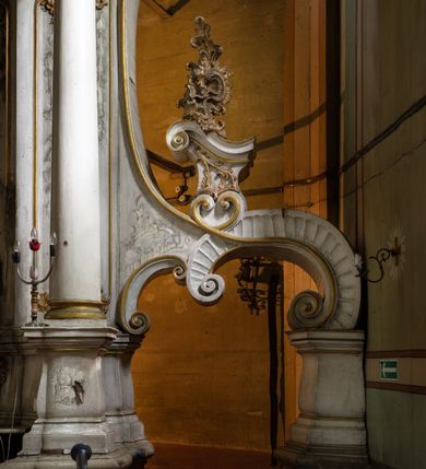 Zdjęcie nr 4: Kamienny ołtarz zamknięto w drewnianej obudowie, nakrywając go drewnianą mensą z portatylem. Stipes ołtarzowy uzyskał kształt prostopadłościanu. Antepedium ujęte zostało złoconą ramą dekorowaną rocaille’m. W jego polu umieszczono haftowaną tkaninę z medalionem w centrum z przedstawieniem w glorii chrystogramu "IHS", flankowany przez wić różaną. Na mensie ołtarza usytuowano tabernakulum w formie stojącego prostopadłościanu o płaskim froncie i wklęsłych bokach pozbawionych cokołu. Drzwiczki wprawiono w płaską niszę zwieńczoną profilowanym łukiem odcinkowym, nadwieszonym. U góry tabernakulum flankuje para sakramentek. Na tabernakulum usytuowano tron eucharystyczny utworzony z dwóch ażurowych spływów wolutowych podtrzymujących baldachim z lambrekinem. Na baldachimie na czerwonej poduszce spoczywa płaska, złocona korona otwarta. Retabulum ołtarzowe wzniesiono na planie półowalu i polichromowano je na biało. Jest to architektoniczna, jednokondygnacyjna, jednoosiowa struktura ze zwieńczeniem. Po bokach flankują ją bramki ustawione na dwustrefowym, tralkowym, wklęsło-wypukłym cokole. Łuki bramek tworzą fantazyjnie wygięte dwie woluty. Kondygnacja została ujęta dwiema parami pilastrów zwróconych delikatnie do wewnątrz oraz silnie wysuniętą do przodu parą kolumn o kapitelach kompozytowych. Podpory dźwigają pełne belkowanie o wklęsło-wypukłej linii. Nad polem środkowym belkowanie jest połączone gzymsem przyjmującym formę łuku falistego i wyłamane na osiach podpór. Kondygnację ujmują spływy wolutowe połączone z łukowato wygiętymi wolutami podtrzymywanymi przez wysokie tralkowe cokoły, tworząc bramki ołtarza. Bramki zwieńczono fantazyjnie wygiętymi impostami podtrzymującymi ornament rocalliowy. W polu środkowym umieszczono wielkoformatowe zdjęcie znajdującego się tu pierwotnie obrazu św. Mikołaja, ujęte profilowaną, złoconą ramą w formie stojącego prostokąta zamkniętego łukiem półkolistym nadwieszonym z uskokiem. Zwieńczenie uzyskało formę leżącego prostokąta flankowanego parą pilastrów zwróconych do wewnątrz oraz parą pilastrów zwróconych na zewnątrz, których trzony przechodzą w spływy wolutowe opadające na belkowanie. Pilastry dźwigają niepełne, wklęsło-wypukłe belkowanie przyjmujące nad polem formę łuku falistego. W polu zwieńczenia znalazło się przedstawienie gołębicy Ducha Świętego z ażurowymi promieniami na tle owalnego okna prezbiterium, ujęte w fantazjną, wklęsło-wypukłą, profilowaną i złoconą ramę. Pilastry kondygnacji ozdobiono kapitelami z rocaille, a ich trzony udekorowano złoconymi, rokokowymi panneau ze zwisami utworzonymi z rocaille, rozet i liści. Na ich tle usytuowano pierwotnie figury św. Kazimierza Królewicza i św. Augustyna (obecnie przeniesione do krypty nowego kościoła). Rzeźby stały na górnej strefie cokołu na wspornikach z rocaille. W przyłuczach i na belkowaniu znalazły się ornamenty roślinne. Także trzony pilastrów zwieńczenia dekorowane są złoconymi zwisami z rocaille i kwiatów. Pole wokół promieni z gołębicą Ducha Świętego również ozdobiono fragmentami rocaille. 