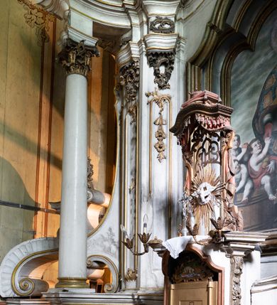 Zdjęcie nr 14: Kamienny ołtarz zamknięto w drewnianej obudowie, nakrywając go drewnianą mensą z portatylem. Stipes ołtarzowy uzyskał kształt prostopadłościanu. Antepedium ujęte zostało złoconą ramą dekorowaną rocaille’m. W jego polu umieszczono haftowaną tkaninę z medalionem w centrum z przedstawieniem w glorii chrystogramu "IHS", flankowany przez wić różaną. Na mensie ołtarza usytuowano tabernakulum w formie stojącego prostopadłościanu o płaskim froncie i wklęsłych bokach pozbawionych cokołu. Drzwiczki wprawiono w płaską niszę zwieńczoną profilowanym łukiem odcinkowym, nadwieszonym. U góry tabernakulum flankuje para sakramentek. Na tabernakulum usytuowano tron eucharystyczny utworzony z dwóch ażurowych spływów wolutowych podtrzymujących baldachim z lambrekinem. Na baldachimie na czerwonej poduszce spoczywa płaska, złocona korona otwarta. Retabulum ołtarzowe wzniesiono na planie półowalu i polichromowano je na biało. Jest to architektoniczna, jednokondygnacyjna, jednoosiowa struktura ze zwieńczeniem. Po bokach flankują ją bramki ustawione na dwustrefowym, tralkowym, wklęsło-wypukłym cokole. Łuki bramek tworzą fantazyjnie wygięte dwie woluty. Kondygnacja została ujęta dwiema parami pilastrów zwróconych delikatnie do wewnątrz oraz silnie wysuniętą do przodu parą kolumn o kapitelach kompozytowych. Podpory dźwigają pełne belkowanie o wklęsło-wypukłej linii. Nad polem środkowym belkowanie jest połączone gzymsem przyjmującym formę łuku falistego i wyłamane na osiach podpór. Kondygnację ujmują spływy wolutowe połączone z łukowato wygiętymi wolutami podtrzymywanymi przez wysokie tralkowe cokoły, tworząc bramki ołtarza. Bramki zwieńczono fantazyjnie wygiętymi impostami podtrzymującymi ornament rocalliowy. W polu środkowym umieszczono wielkoformatowe zdjęcie znajdującego się tu pierwotnie obrazu św. Mikołaja, ujęte profilowaną, złoconą ramą w formie stojącego prostokąta zamkniętego łukiem półkolistym nadwieszonym z uskokiem. Zwieńczenie uzyskało formę leżącego prostokąta flankowanego parą pilastrów zwróconych do wewnątrz oraz parą pilastrów zwróconych na zewnątrz, których trzony przechodzą w spływy wolutowe opadające na belkowanie. Pilastry dźwigają niepełne, wklęsło-wypukłe belkowanie przyjmujące nad polem formę łuku falistego. W polu zwieńczenia znalazło się przedstawienie gołębicy Ducha Świętego z ażurowymi promieniami na tle owalnego okna prezbiterium, ujęte w fantazjną, wklęsło-wypukłą, profilowaną i złoconą ramę. Pilastry kondygnacji ozdobiono kapitelami z rocaille, a ich trzony udekorowano złoconymi, rokokowymi panneau ze zwisami utworzonymi z rocaille, rozet i liści. Na ich tle usytuowano pierwotnie figury św. Kazimierza Królewicza i św. Augustyna (obecnie przeniesione do krypty nowego kościoła). Rzeźby stały na górnej strefie cokołu na wspornikach z rocaille. W przyłuczach i na belkowaniu znalazły się ornamenty roślinne. Także trzony pilastrów zwieńczenia dekorowane są złoconymi zwisami z rocaille i kwiatów. Pole wokół promieni z gołębicą Ducha Świętego również ozdobiono fragmentami rocaille. 