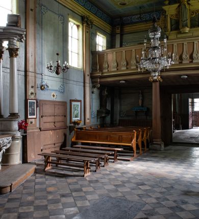 Zdjęcie nr 12: Drewniany kościół pw. św. Mikołaja w Polance jest budowlą orientowaną. Do prostokątnej nawy przylega węższe, wydłużone, trójbocznie zamknięte prezbiterium z piętrową zakrystią na osi o poligonalnym zamknięciu. Do zakrystii przylega od południa kruchta. Fragment północnej ściany prezbiterium jest murowany. Od zachodu do nawy dobudowano czworoboczną wieżę poprzedzoną przedsionkiem, który również dostawiono do nawy od południa. Prezbiterium i nawę wzniesiono na zrąb, natomiast wieżę w konstrukcji słupowej. Wieża na rzucie kwadratu posiada dwie kondygnacje. Dolna jest tej samej szerokości co nawa, górna natomiast znacznie węższa. Jej masyw podzielono smukłymi pilastrami i lizenami, zaś poniżej profilowanego gzymsu koronującego umieszczono wyrzynaną w desce koronkę. Kondygnacje oddzielono daszkiem gontowym zlewającym się po bokach z dachem kościoła. Wieżę nakryto kopulastym hełmem blaszanym, zwieńczonym blaszanym obeliskiem z krzyżem. Prezbiterium i nawę nakryto eternitowym dachem siodłowym podkreślonym profilowanym gzymsem. Nad nawą w części wschodniej zainstalowano wieżyczkę na sygnaturkę w formie wielobocznego graniastosłupa przykrytego cebulastą banią z krzyżem. Dokoła kościoła założono daszek gontowy chroniący podwaliny. Pod kościołem urządzono kryptę grzebalną. Wnętrze kościoła artykułowano za pomocą pilastrów i gzymsu. Całość nakryto stropem z fasetą. Otwór tęczowy uzyskał kształt trójlistnego, ściętego łuku, wspartego na filarkach ustawionych na profilowanej belce zawieszonej na kroksztynach. W części zachodniej nawy urządzono chór muzyczny, wsparty na dwóch kolumnach jońskich z przedpiersiem rozczłonkowanym balasami. We wnętrzu ustawiono ołtarz główny i dwa ołtarze boczne oraz ambonę, a na chórze muzycznym zainstalowano prospekt organowy.