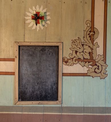 Zdjęcie nr 2: Polichromia malarska we wnętrzu drewnianego kościoła parafialnego w Polance Wielkiej zachowała się w prezbiterium, nawie oraz w przedsionku urządzonym w przyziemiu wieży. W prezbiterium i nawie zastosowano podział ramowy uwzględniający drewniane pilastry artykułujące ściany, gzyms oraz fasetę. Na ścianach pomiędzy pilastrami wprowadzono czworoboczne panneau o narożach utworzonych w stylizowanych formach nawiązujących do motywów roślinno-kwiatowych. Fasetę udekorowano rytmicznie rozłożonym ornamentem z motywów kwiatowych w formie dekoracyjnego fryzu, korespondującego z ornamentalną bordiurą poprowadzoną wzdłuż krawędzi płaskiego stropu. Na stropie w prezbiterium wyobrażono Trójcę Świętą, natomiast w nawie Matkę Boską Różańcową otoczoną przez tajemnice różańcowe w medalionach. Trójcę Świętą wyobrażono na obłokach na niebieskim tle usianym złotymi gwiazdkami. Frontalnie ujęty Bóg Ojciec został ukazany jako siwobrody starzec o bujnych siwych włosach. W prawej ręce trzyma złote berło, lewą zaś wyciąga przed siebie. Bóg Ojciec stopy wspiera o niebieski glob wyłaniający się z obłoków poniżej. Jego twarz charakteryzuje jasna karnacja, szeroko otwarte oczy, wydatny nos i usta. Głowę otacza długa, siwa broda i podobne włosy. Obok Boga Ojca z lewej strony siedzi Chrystus, który obie nogi wspiera na globie. Zbawiciel prawą ręką podtrzymuje oparty o ramię drewniany krzyż, lewą zaś ma odsuniętą od siebie. Jezus został przedstawiony w trzech czwartych w prawo. Jest to dojrzały mężczyzna o pociągłej twarzy okolonej brązowymi, długimi włosami spływającymi na ramiona oraz brodą. Ma jasną karnację, prosty nos i wydatne usta. Nagi tors okrywa bladoszkarłatny płaszcz odsłaniający prawą nogę. Na dłoniach i stopach widoczne są rany po gwoździach. Głowę Jezusa okala świetlisty nimb. Powyżej obu postaci na tle rozjaśnionego nieba unosi się gołębica Ducha Świętego w promienistym otoku. Polichromia stropu w nawie została zakomponowana w oparciu o zmniejszające się koncentryczne okręgi wpisane w dekoracjne panneau o narożach utworzonych ze stylizowanych form nawiązujących do motywów roślinno-kwiatowych. W środkowym okręgu ukazano zasiadającą na obłokach Matkę Boską z Dzieciątkiem otoczoną okrągłym obramieniem utworzonym z wielobarwnego ornamentu z motywem naprzemiennie ułożonych lancetowatych liści-łusek. Madonna została przedstawiona frontalnie, z prawą ręką przyłożoną do piersi, lewą zaś obejmuje Dzieciątko Jezus siedzące na globie ułożonym na kolanach matki podtrzymywanym przez anioła. Maryja ma pociągłą twarz o wydatnym nosie i wargach. Jej spojrzenie półprzymkniętych oczu skierowane jest pokornie ku dołowi. Głowę Marii otaczają, brązowe, falujące włosy spływające na ramiona. Madonna ubrana jest w czerwoną suknię przewiązaną złocistą szarfą w pasie i niebieski płaszcz lamowany złotą taśmą, spięty pod szyją z lewą połą osłaniającą jej kolana. Lewą stopę Matka Boska wspiera o wystający spod płaszcza poniżej złoty półksiężyc. Siedzące na globie Dzieciątko ujęte zostało w trzech czwartych w lewo. Mały Jezus prawą ręką nakłada matce złotą koronę, w lewej zaś trzyma lilię. Zbawiciel ma owalną twarz o jasnej karnacjim i rumianych policzkach z drobnym nosem i ustami. Jego głowę okalają jasne loki. Ubrany jest w jasną przepaskę przerzuconą przez prawe ramię oraz biodra. Głowy obu postaci otaczają świetliste promienie. Z prawej strony Marii stoi ujęty w trzech czwarty w prawo anioł, z twarzą zwróconą w stronę widza, podtrzymujący oburącz niebieski glob, na którym siedzi Jezus. Uskrzydlony anioł o jasnobrązowych włosach ubrany jest w zieloną tunikę przewiązaną w pasie. Za Madonną z lewej strony widoczny jest asystujący drugi anioł w cynobrowej tunice i jasnych włosach spływających na ramiona. Pod półksiężycem na tle obłoków widoczne są trzy uskrzydlone główki anielskie. Wokół Matki Boskiej rozmieszczono owalne medaliony z blachy miedzianej malowane w piętnaście tajemnic Różańca świętego. Ich narracja rozpoczyna się nad głową Madonny. Każda ze scen oparta jest o kilkuosobową kompozycję. Idąc w lewo namalowano: Zwiastowanie Marii, Nawiedzenie św. Elżbiety, Boże Narodzenie, Ofiarowanie w świątyni, Znalezienie w świątyni, Modlitwa w Ogrójcu, Biczowanie, Cierniem ukoronowanie, Niesienie krzyża, Ukrzyżowanie, Zmartwychwstanie, Wniebowstąpienie, Zesłanie Ducha Świętego, Wniebowzięcie Marii, Ukoronowanie Marii w niebie. Pomiędzy medalionami namalowano czerwone róże. Całoś obramienia ornamentalny okrąg. Dekorację w przedsionku utworzono za pomocą iluzjonistycznie malowanych cynobrowych kolumn o korynckich kapitelach ustawionych na wysokich postumentach, podtrzymujących belkowanie. Pomiędzy kolumnami ściany są gładkie, bladoniebieskie. Strop ozdobiono obramieniem w formie czworobocznego panneau o narożach utworzonych ze stylizowanych form nawiązujących do motywów roślinno-kwiatowych. 