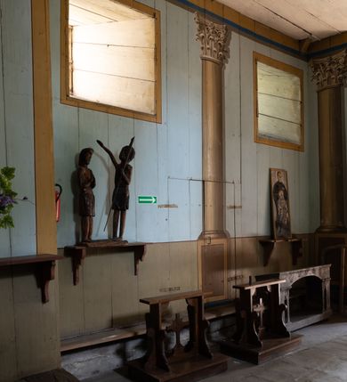 Zdjęcie nr 15: Polichromia malarska we wnętrzu drewnianego kościoła parafialnego w Polance Wielkiej zachowała się w prezbiterium, nawie oraz w przedsionku urządzonym w przyziemiu wieży. W prezbiterium i nawie zastosowano podział ramowy uwzględniający drewniane pilastry artykułujące ściany, gzyms oraz fasetę. Na ścianach pomiędzy pilastrami wprowadzono czworoboczne panneau o narożach utworzonych w stylizowanych formach nawiązujących do motywów roślinno-kwiatowych. Fasetę udekorowano rytmicznie rozłożonym ornamentem z motywów kwiatowych w formie dekoracyjnego fryzu, korespondującego z ornamentalną bordiurą poprowadzoną wzdłuż krawędzi płaskiego stropu. Na stropie w prezbiterium wyobrażono Trójcę Świętą, natomiast w nawie Matkę Boską Różańcową otoczoną przez tajemnice różańcowe w medalionach. Trójcę Świętą wyobrażono na obłokach na niebieskim tle usianym złotymi gwiazdkami. Frontalnie ujęty Bóg Ojciec został ukazany jako siwobrody starzec o bujnych siwych włosach. W prawej ręce trzyma złote berło, lewą zaś wyciąga przed siebie. Bóg Ojciec stopy wspiera o niebieski glob wyłaniający się z obłoków poniżej. Jego twarz charakteryzuje jasna karnacja, szeroko otwarte oczy, wydatny nos i usta. Głowę otacza długa, siwa broda i podobne włosy. Obok Boga Ojca z lewej strony siedzi Chrystus, który obie nogi wspiera na globie. Zbawiciel prawą ręką podtrzymuje oparty o ramię drewniany krzyż, lewą zaś ma odsuniętą od siebie. Jezus został przedstawiony w trzech czwartych w prawo. Jest to dojrzały mężczyzna o pociągłej twarzy okolonej brązowymi, długimi włosami spływającymi na ramiona oraz brodą. Ma jasną karnację, prosty nos i wydatne usta. Nagi tors okrywa bladoszkarłatny płaszcz odsłaniający prawą nogę. Na dłoniach i stopach widoczne są rany po gwoździach. Głowę Jezusa okala świetlisty nimb. Powyżej obu postaci na tle rozjaśnionego nieba unosi się gołębica Ducha Świętego w promienistym otoku. Polichromia stropu w nawie została zakomponowana w oparciu o zmniejszające się koncentryczne okręgi wpisane w dekoracjne panneau o narożach utworzonych ze stylizowanych form nawiązujących do motywów roślinno-kwiatowych. W środkowym okręgu ukazano zasiadającą na obłokach Matkę Boską z Dzieciątkiem otoczoną okrągłym obramieniem utworzonym z wielobarwnego ornamentu z motywem naprzemiennie ułożonych lancetowatych liści-łusek. Madonna została przedstawiona frontalnie, z prawą ręką przyłożoną do piersi, lewą zaś obejmuje Dzieciątko Jezus siedzące na globie ułożonym na kolanach matki podtrzymywanym przez anioła. Maryja ma pociągłą twarz o wydatnym nosie i wargach. Jej spojrzenie półprzymkniętych oczu skierowane jest pokornie ku dołowi. Głowę Marii otaczają, brązowe, falujące włosy spływające na ramiona. Madonna ubrana jest w czerwoną suknię przewiązaną złocistą szarfą w pasie i niebieski płaszcz lamowany złotą taśmą, spięty pod szyją z lewą połą osłaniającą jej kolana. Lewą stopę Matka Boska wspiera o wystający spod płaszcza poniżej złoty półksiężyc. Siedzące na globie Dzieciątko ujęte zostało w trzech czwartych w lewo. Mały Jezus prawą ręką nakłada matce złotą koronę, w lewej zaś trzyma lilię. Zbawiciel ma owalną twarz o jasnej karnacjim i rumianych policzkach z drobnym nosem i ustami. Jego głowę okalają jasne loki. Ubrany jest w jasną przepaskę przerzuconą przez prawe ramię oraz biodra. Głowy obu postaci otaczają świetliste promienie. Z prawej strony Marii stoi ujęty w trzech czwarty w prawo anioł, z twarzą zwróconą w stronę widza, podtrzymujący oburącz niebieski glob, na którym siedzi Jezus. Uskrzydlony anioł o jasnobrązowych włosach ubrany jest w zieloną tunikę przewiązaną w pasie. Za Madonną z lewej strony widoczny jest asystujący drugi anioł w cynobrowej tunice i jasnych włosach spływających na ramiona. Pod półksiężycem na tle obłoków widoczne są trzy uskrzydlone główki anielskie. Wokół Matki Boskiej rozmieszczono owalne medaliony z blachy miedzianej malowane w piętnaście tajemnic Różańca świętego. Ich narracja rozpoczyna się nad głową Madonny. Każda ze scen oparta jest o kilkuosobową kompozycję. Idąc w lewo namalowano: Zwiastowanie Marii, Nawiedzenie św. Elżbiety, Boże Narodzenie, Ofiarowanie w świątyni, Znalezienie w świątyni, Modlitwa w Ogrójcu, Biczowanie, Cierniem ukoronowanie, Niesienie krzyża, Ukrzyżowanie, Zmartwychwstanie, Wniebowstąpienie, Zesłanie Ducha Świętego, Wniebowzięcie Marii, Ukoronowanie Marii w niebie. Pomiędzy medalionami namalowano czerwone róże. Całoś obramienia ornamentalny okrąg. Dekorację w przedsionku utworzono za pomocą iluzjonistycznie malowanych cynobrowych kolumn o korynckich kapitelach ustawionych na wysokich postumentach, podtrzymujących belkowanie. Pomiędzy kolumnami ściany są gładkie, bladoniebieskie. Strop ozdobiono obramieniem w formie czworobocznego panneau o narożach utworzonych ze stylizowanych form nawiązujących do motywów roślinno-kwiatowych. 