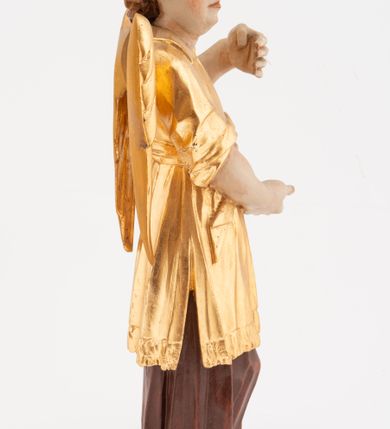 Zdjęcie nr 4: Figura pełnoplastyczna, ustawiona na niskiej podstawie. Postać ukazana frontalnie, z głową skierowaną w prawo, w kontrapoście, z obiema rękami uniesionymi z przodu, jedną wysoko, drugą nisko. Twarz ma podłużną, z drugim podbródkiem, o dużych, migdałowatych i opadających oczach, wąskim nosie i ustach, okoloną krótkimi, kręconymi włosami, układającymi się w czub nad czołem. Anioł ubrany jest w ciemnoczerwoną suknię spodnią i złoconą suknię wierzchnią, sięgającą kolan, z podwiniętymi rękawami, przepasaną w talii szarfą. Skrzydła ma złocone, złożone na plecach. Polichromia ciała naturalistyczna. 