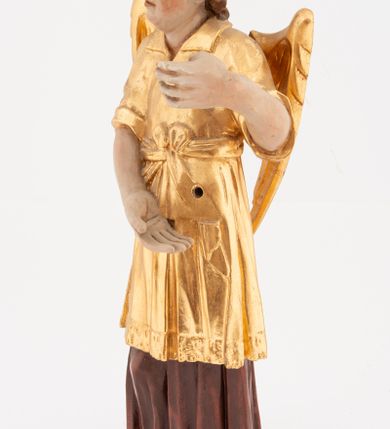 Zdjęcie nr 2: Figura pełnoplastyczna, ustawiona na niskiej podstawie. Postać ukazana frontalnie, z głową skierowaną w prawo, w kontrapoście, z obiema rękami uniesionymi z przodu, jedną wysoko, drugą nisko. Twarz ma podłużną, z drugim podbródkiem, o dużych, migdałowatych i opadających oczach, wąskim nosie i ustach, okoloną krótkimi, kręconymi włosami, układającymi się w czub nad czołem. Anioł ubrany jest w ciemnoczerwoną suknię spodnią i złoconą suknię wierzchnią, sięgającą kolan, z podwiniętymi rękawami, przepasaną w talii szarfą. Skrzydła ma złocone, złożone na plecach. Polichromia ciała naturalistyczna. 